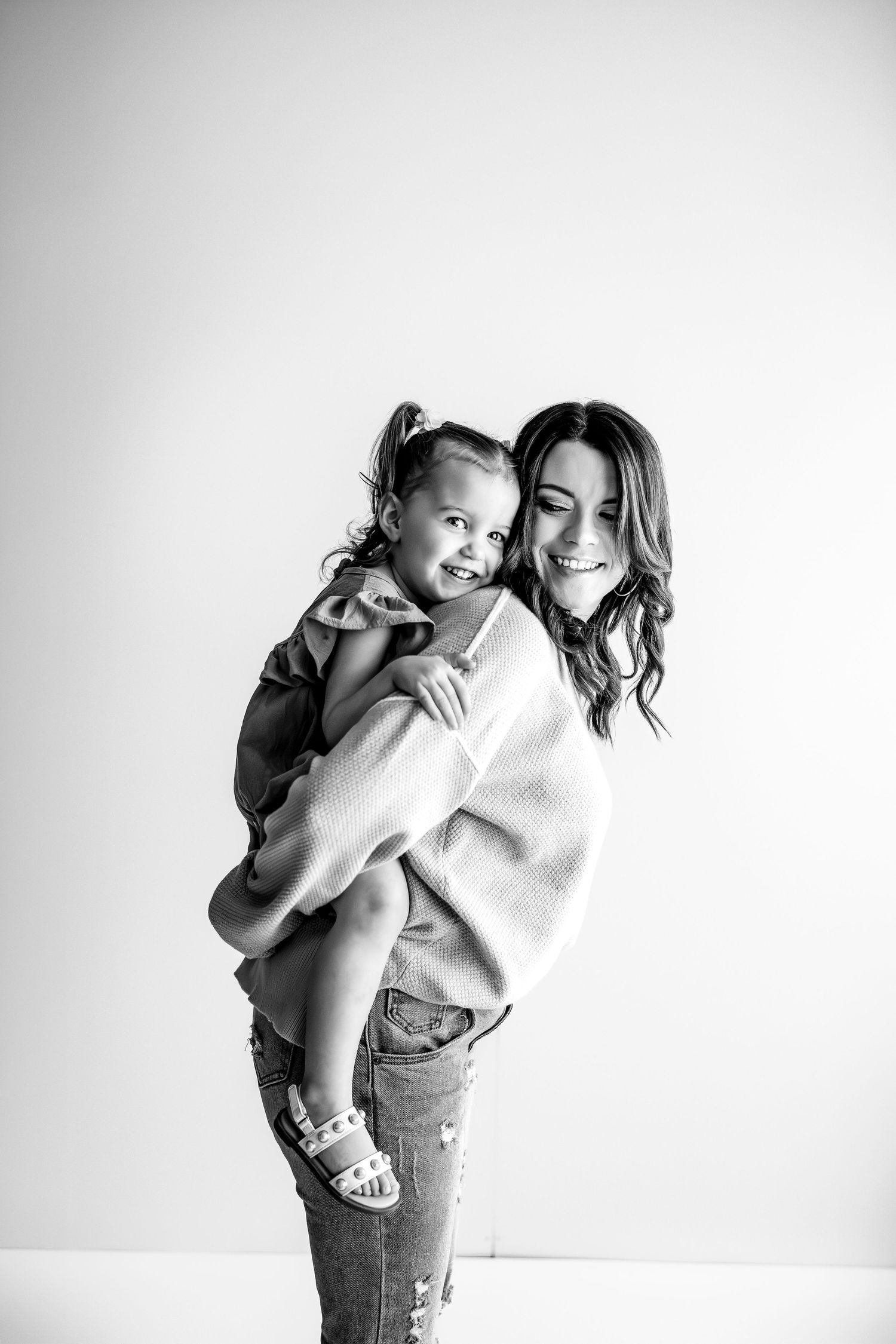 Black and white photo of a smiling woman carrying a happy toddler girl on her back in a bright studio.