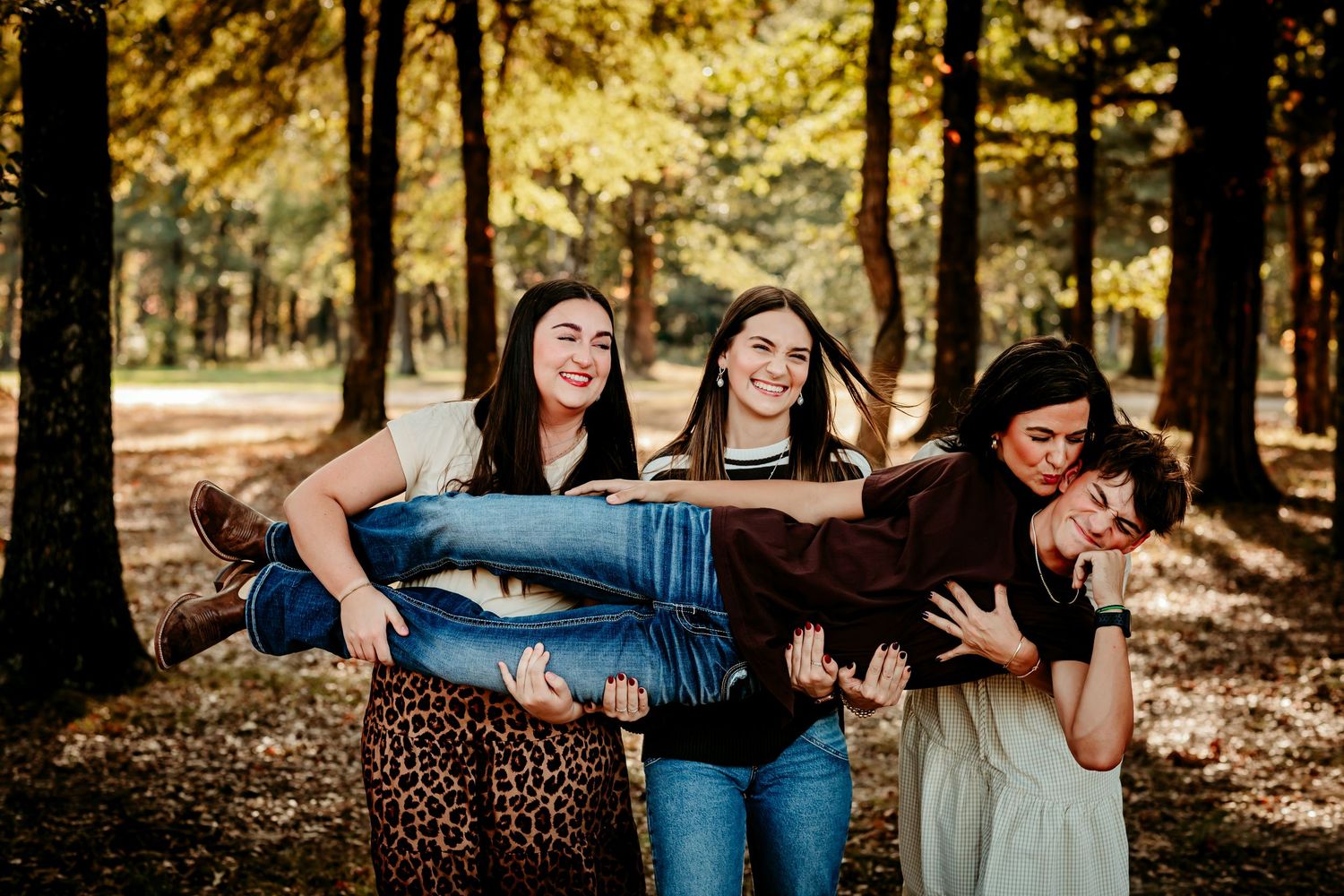 Four young women laughing and playing in an autumn forest, two carrying their friends in a fun outdoor photo session.