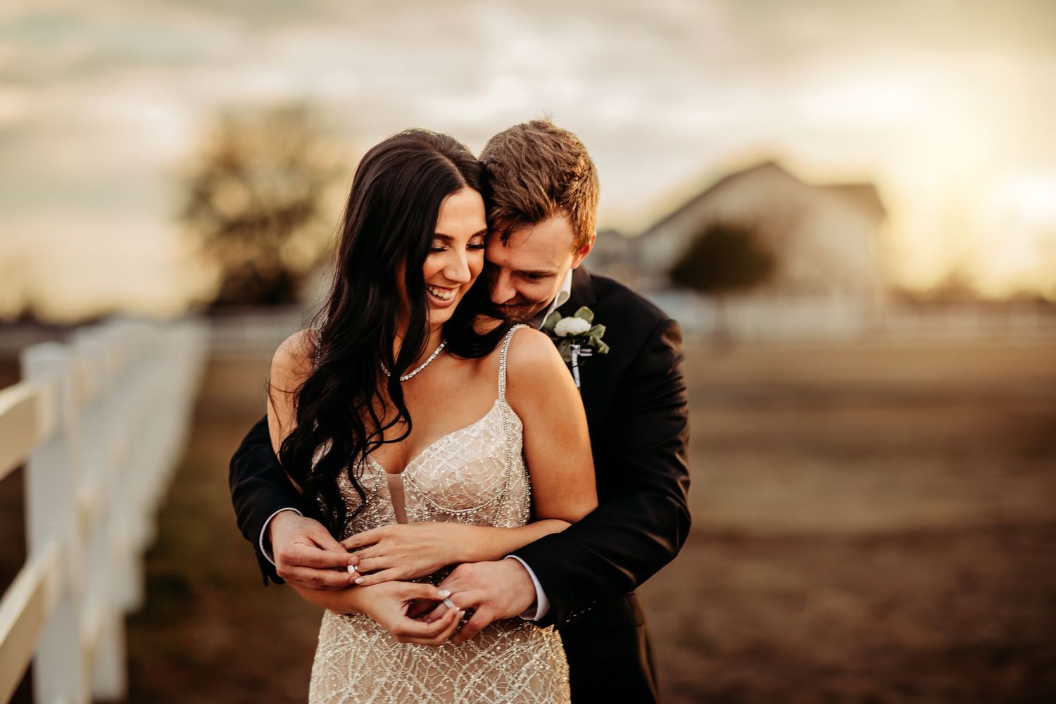 A bride and groom share a romantic embrace at golden hour on a rustic farm, with the bride in a beaded gown.