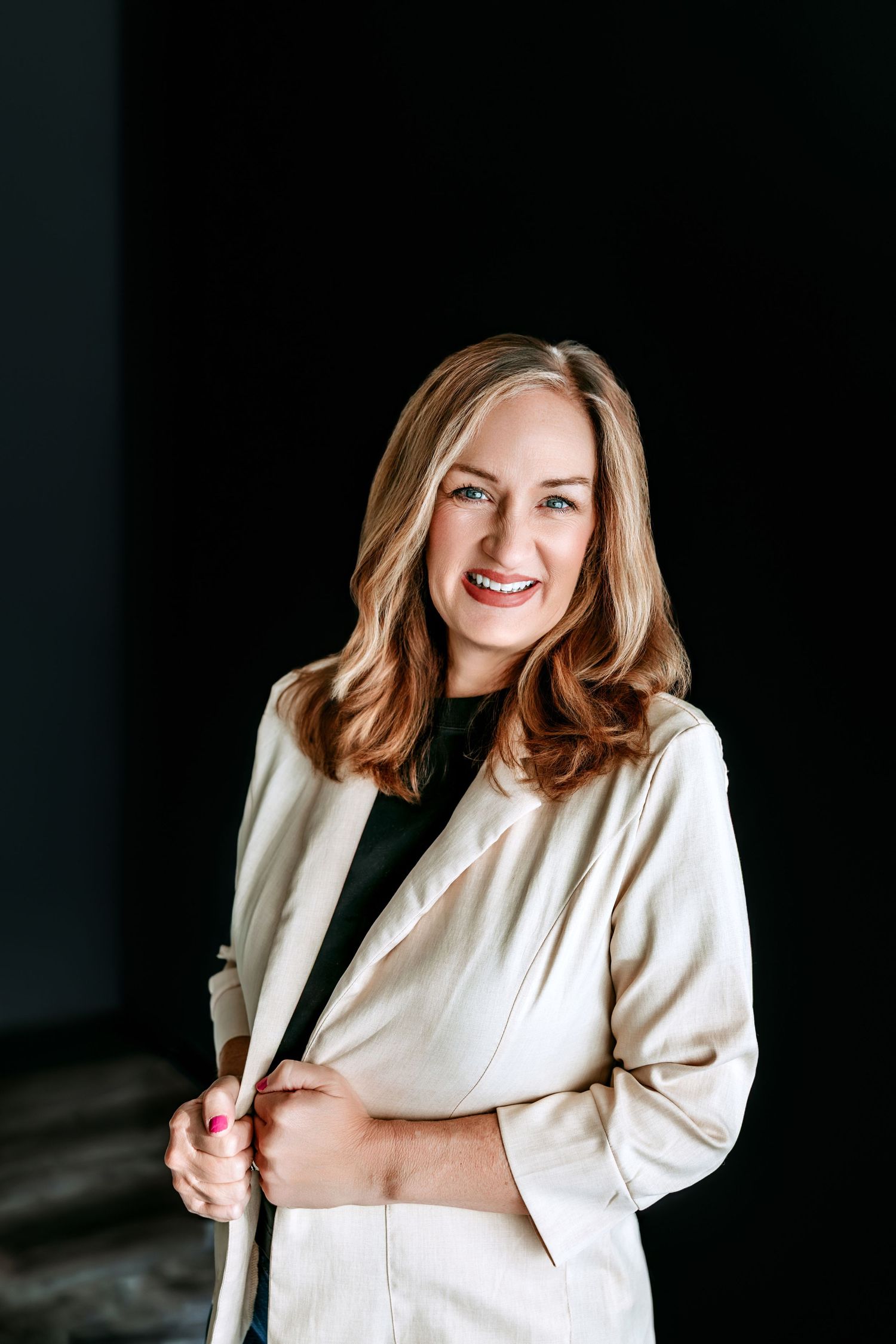 Professional woman in white blazer smiling confidently against dark background, business portrait photography.