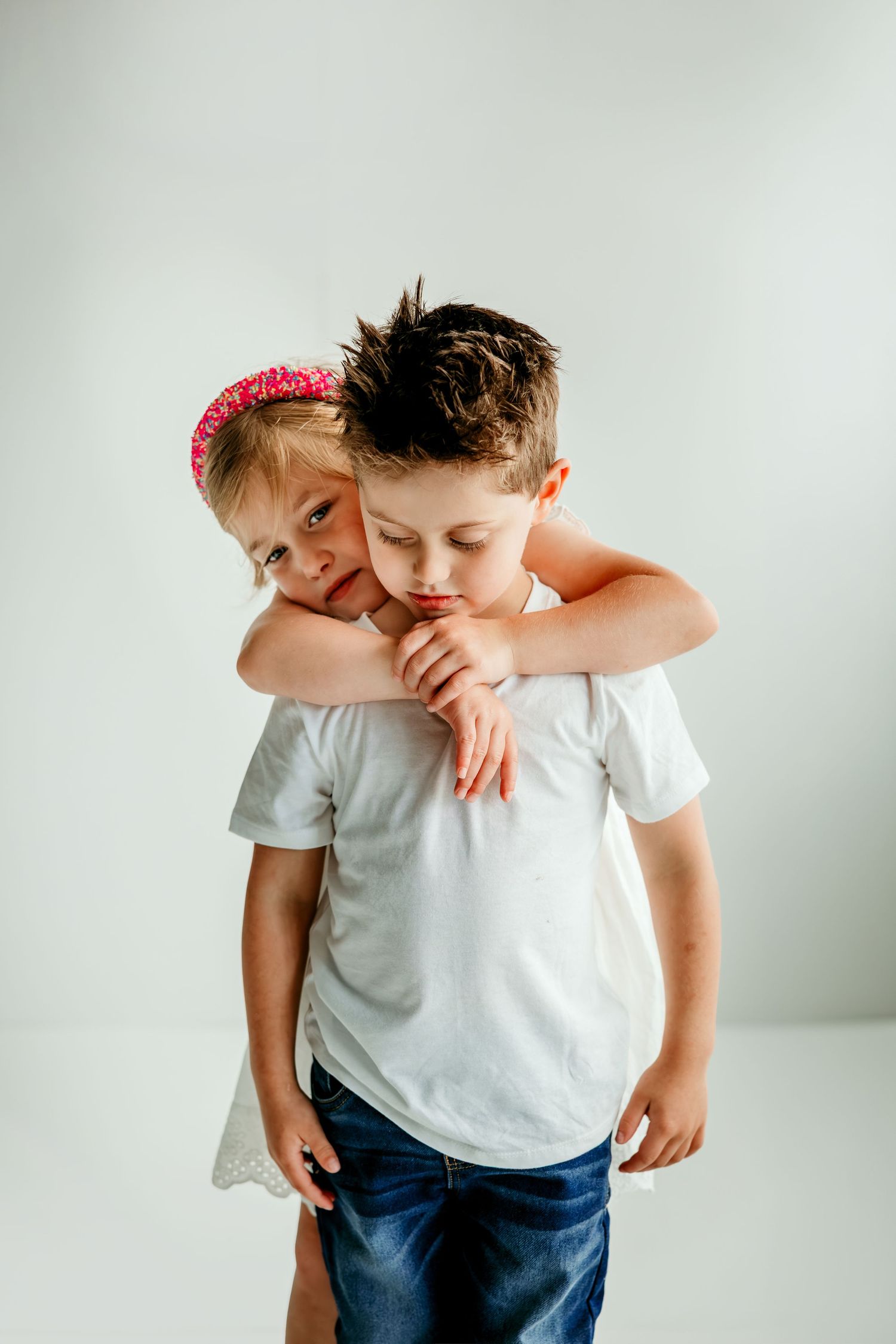 Two young siblings hugging, girl in pink headband embracing boy in white t-shirt against white background.