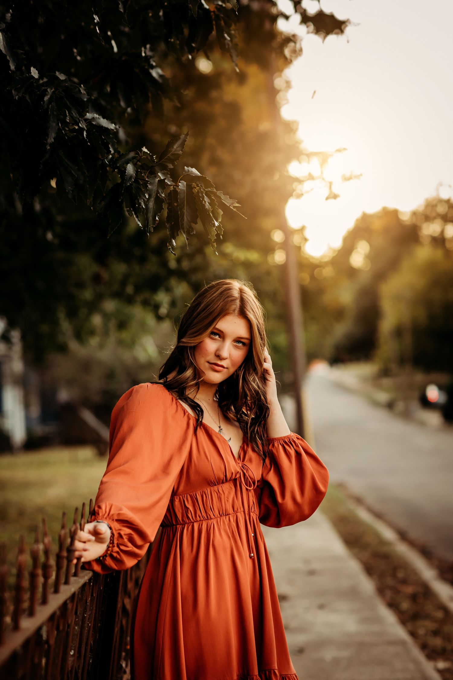 Young woman in rust orange boho dress poses by iron fence at golden hour sunset on tree-lined street.