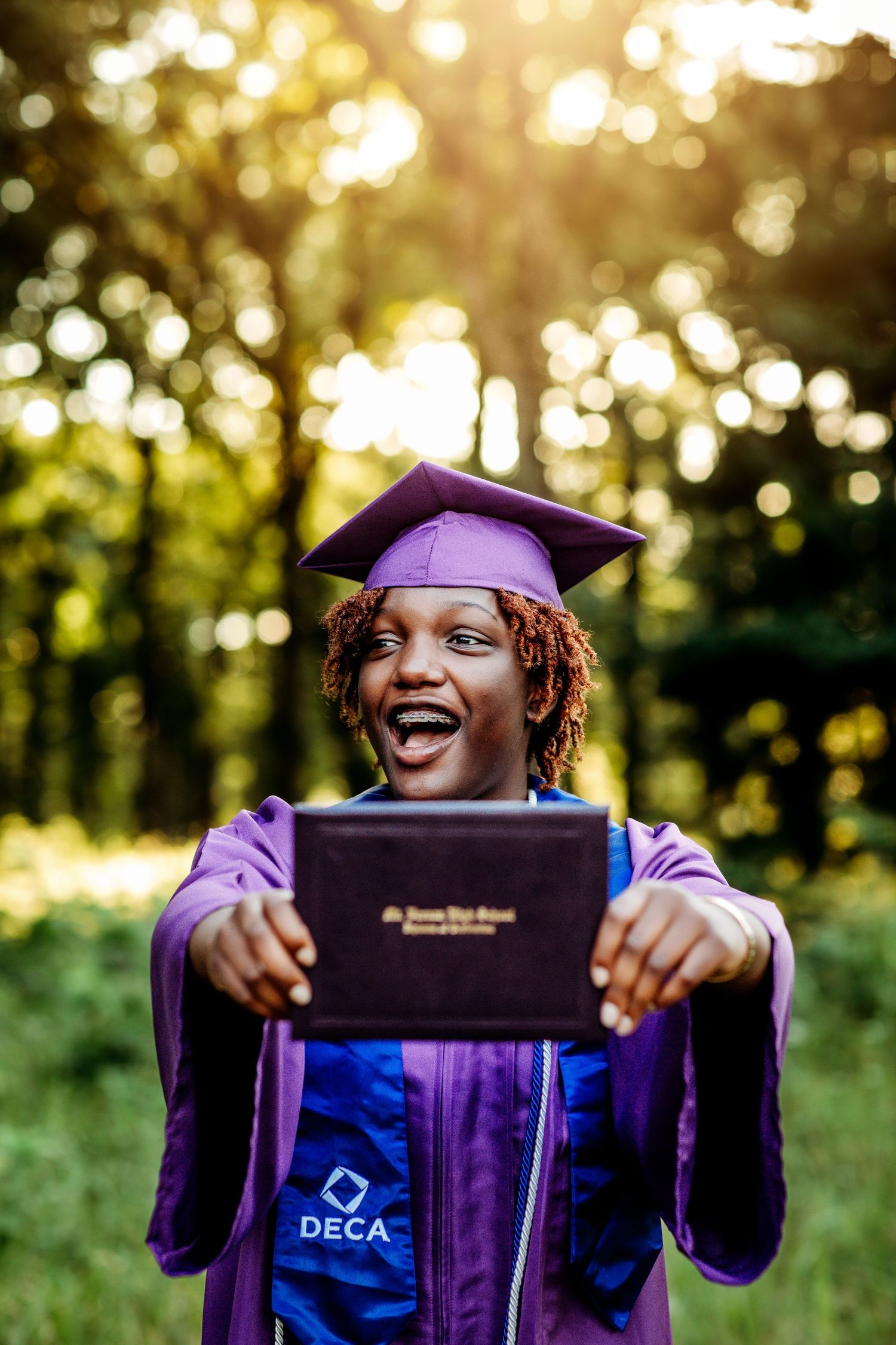 Smiling graduate in purple cap and gown holds diploma outdoors, wearing DECA sash, with sunlit trees in background.