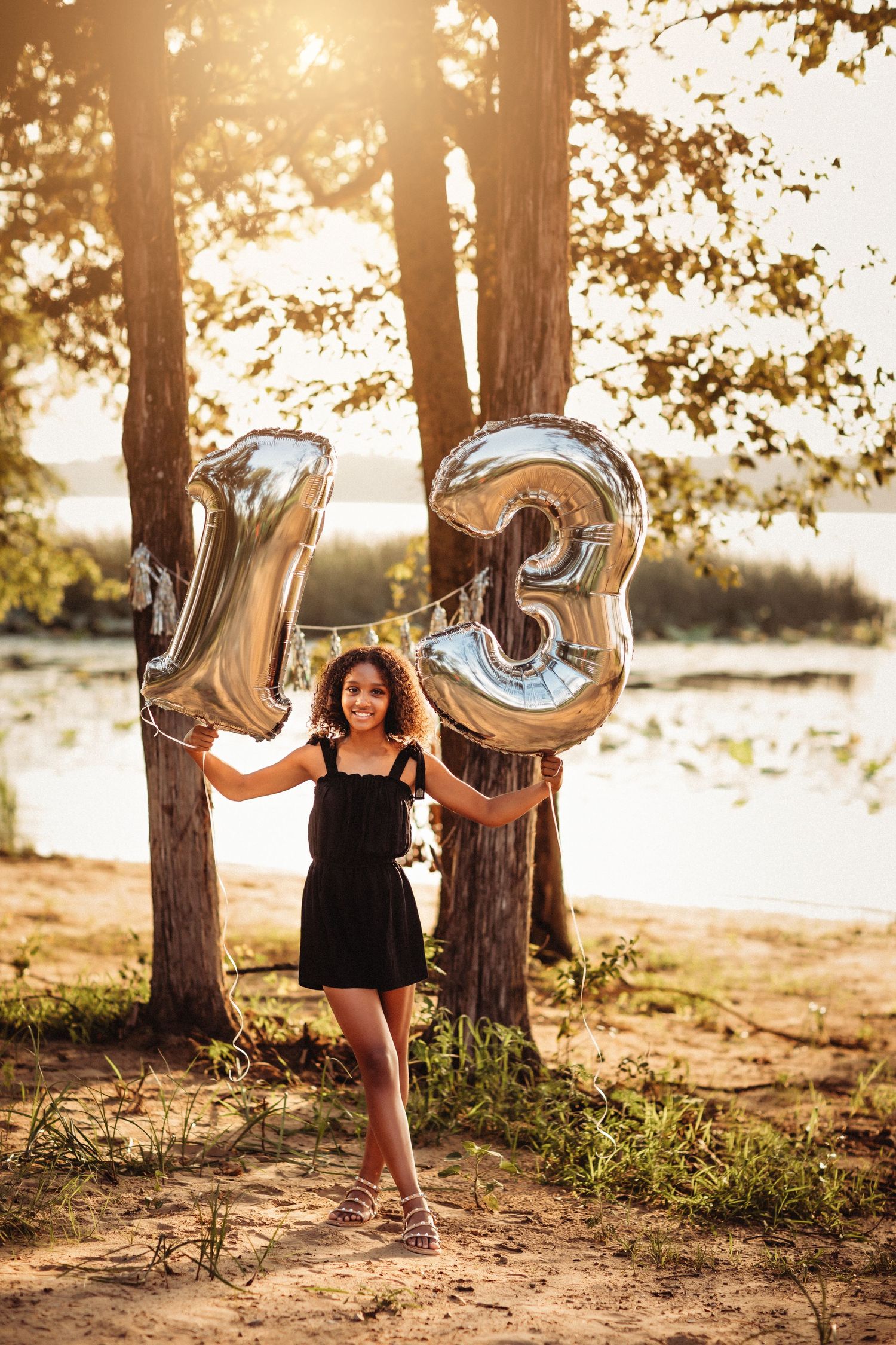 A smiling girl in a black dress holds silver number 13 balloons outdoors near trees and a lake at sunset.