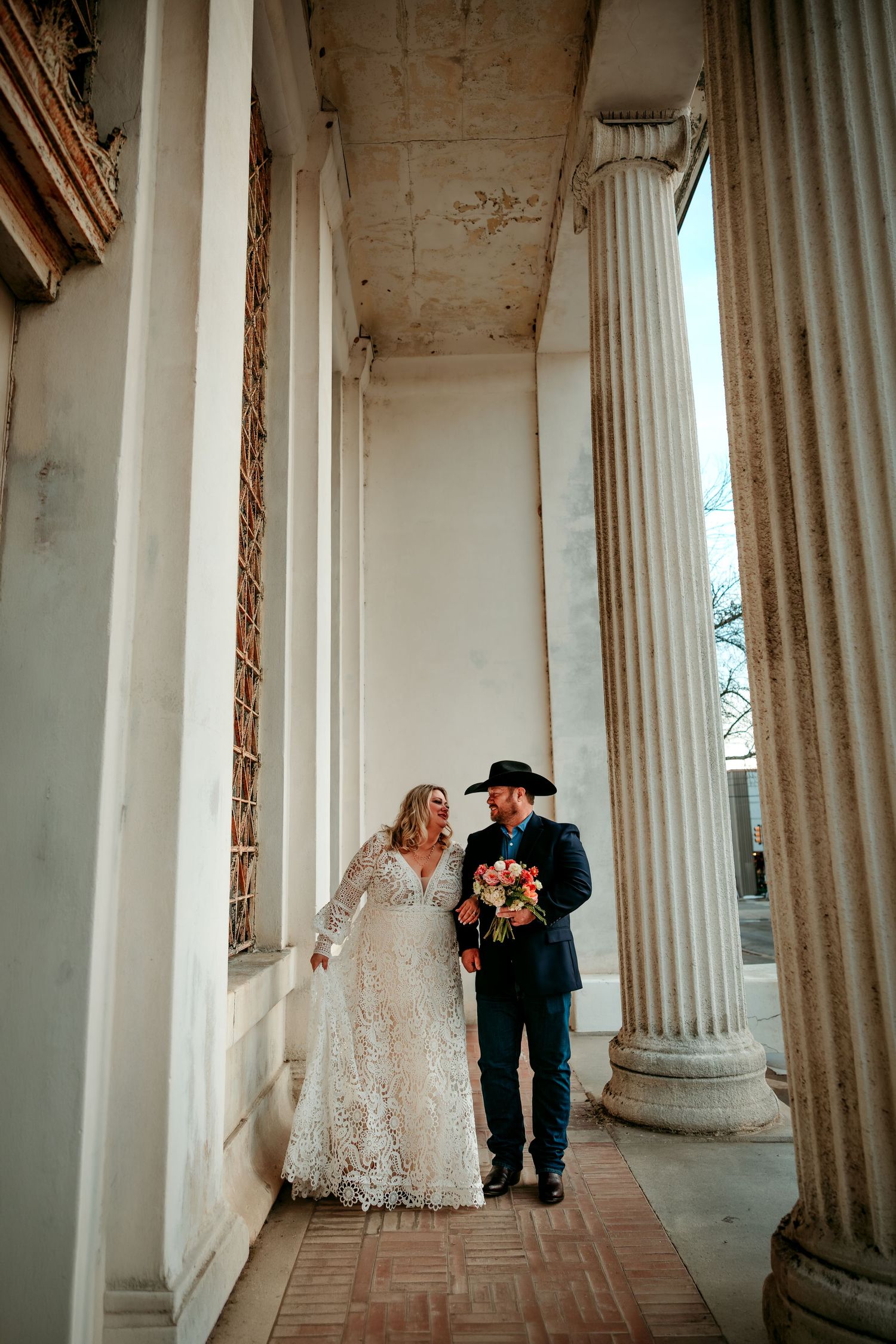 Bride and groom walk among grand white columns, she in lace gown, he in dark suit and cowboy hat, holding colorful bouquet.