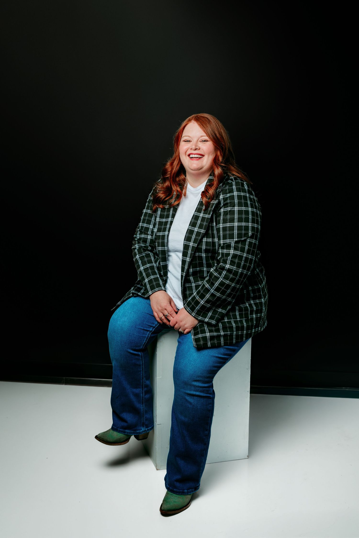 Smiling redhead woman in plaid blazer and jeans sits on white cube against dark background.