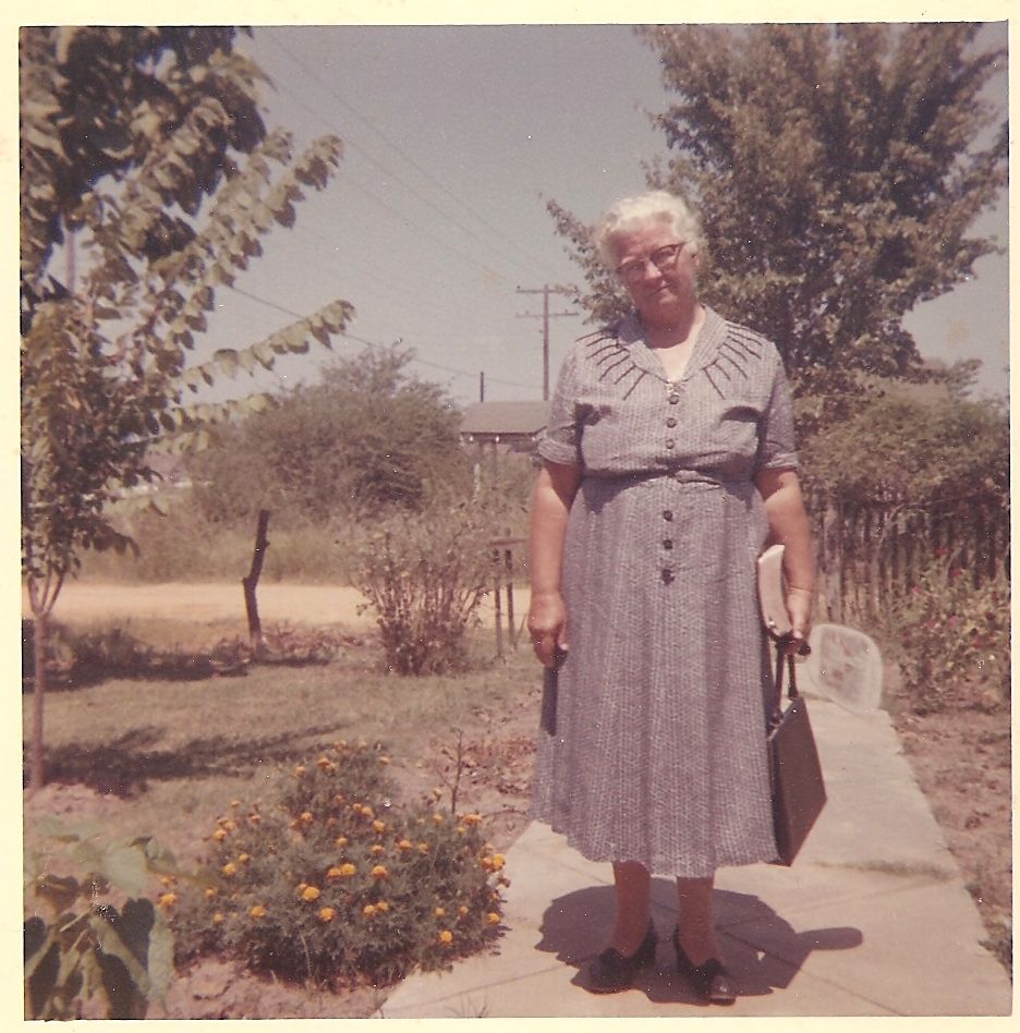 Elderly woman in gray dress holding purse stands on garden path with marigolds and trees in vintage color photo.