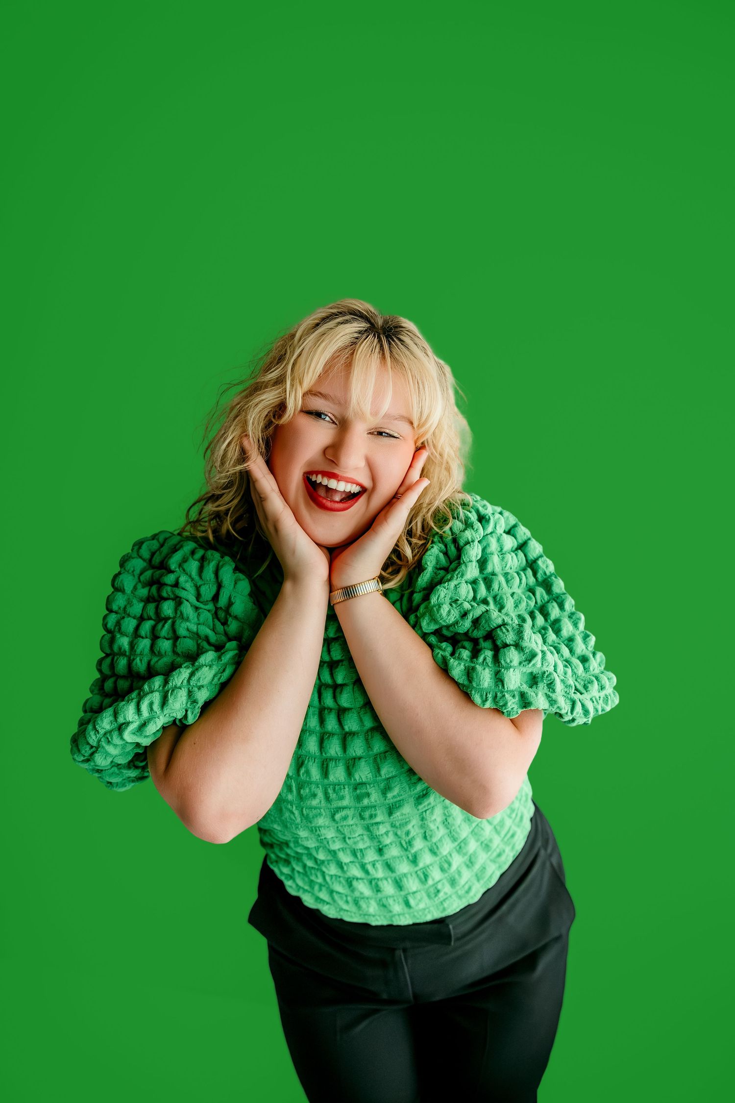 Smiling blonde woman in green chunky knit top poses with hands on cheeks against bright green background.