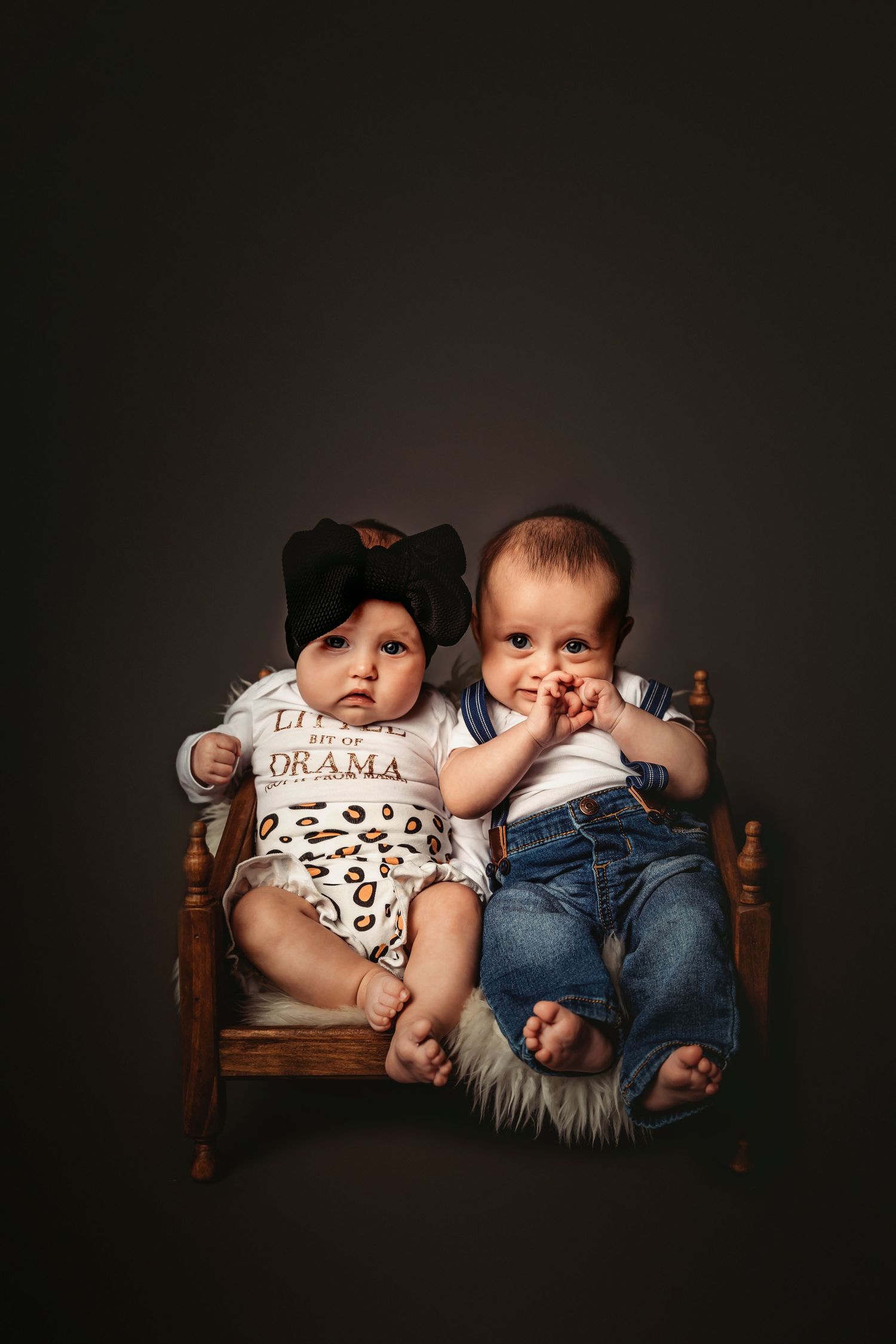 Two babies in casual outfits lying together on a wooden bench against a dark background in a studio setting.