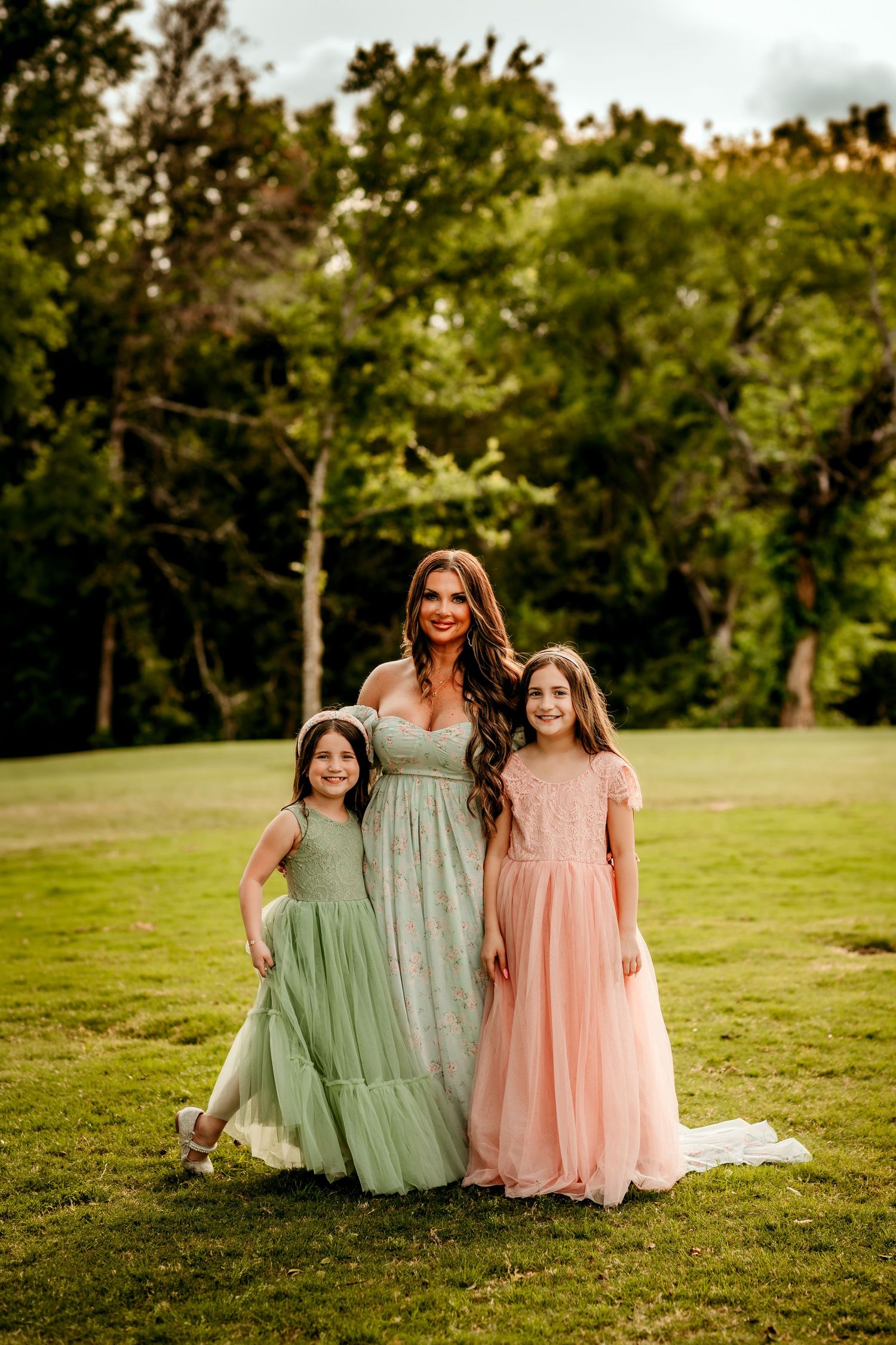 A woman and two girls in elegant green and pink tulle gowns pose together on a lush green lawn surrounded by trees.