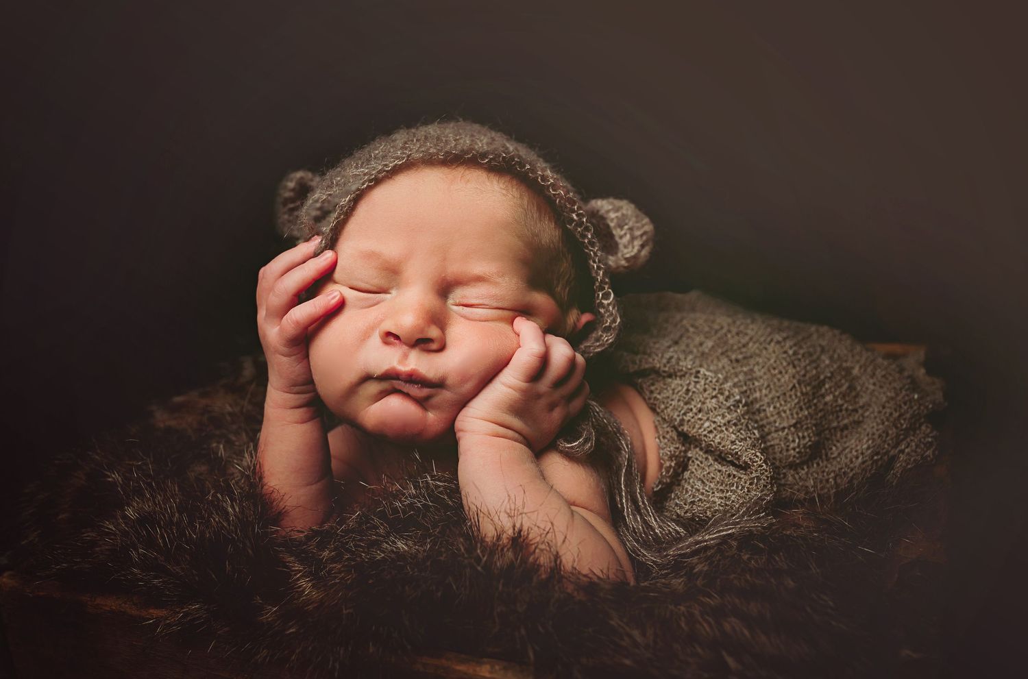 Sleeping newborn baby wearing a knit bear ear hat, posed with hands under chin in a cozy brown nest.