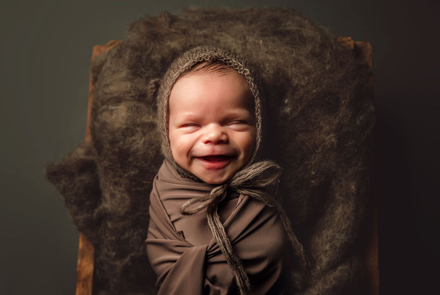Smiling newborn baby wrapped in brown swaddle wearing knit bonnet, posed on rustic wooden prop with soft wool backdrop.