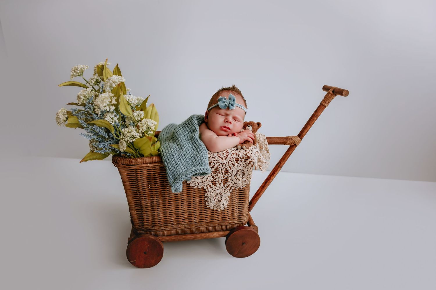 Newborn baby sleeping in a rustic wicker cart with flowers and a blue knit blanket, wearing a floral headband.
