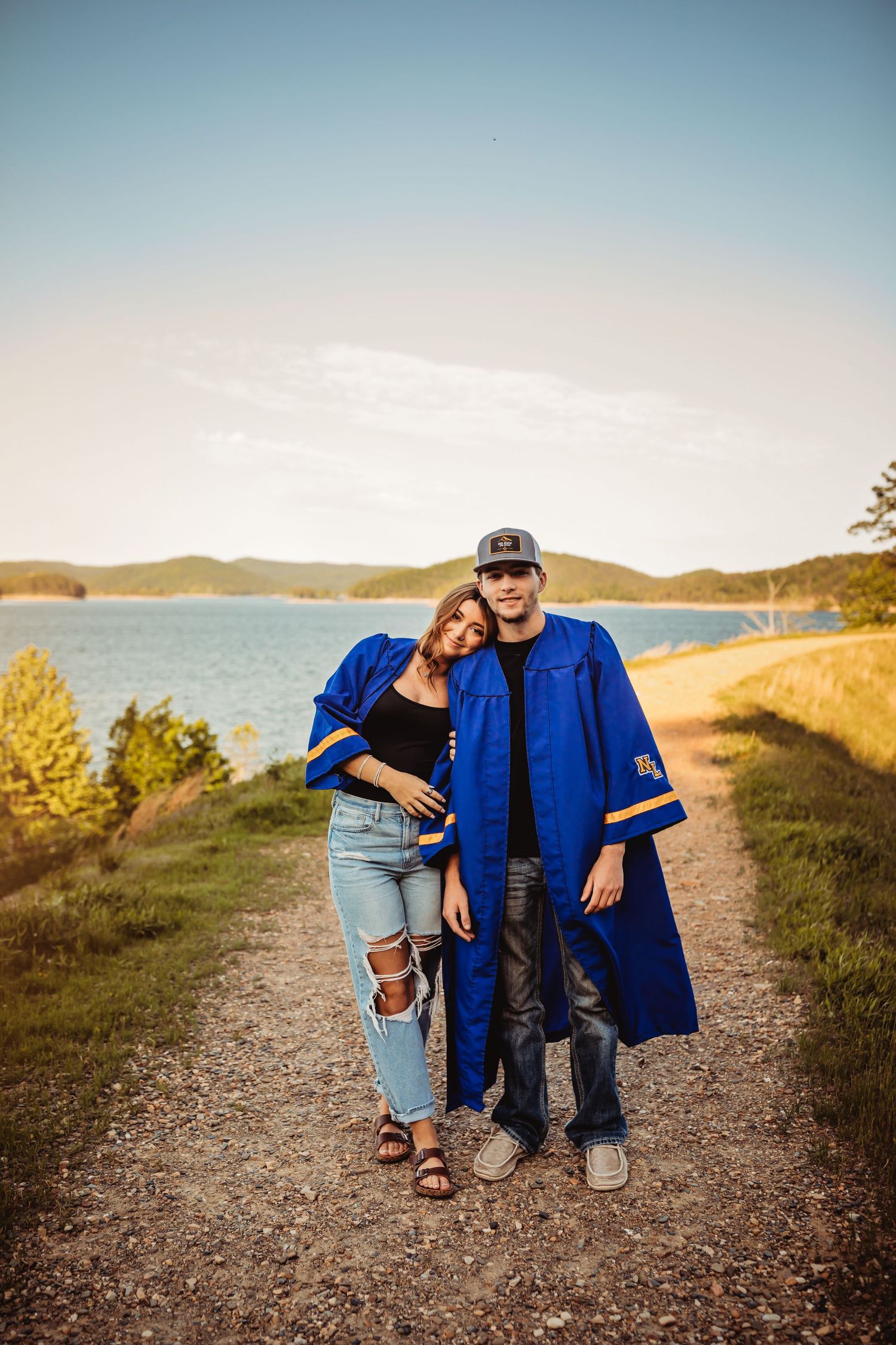 Two graduates in blue caps and gowns pose on a scenic lakeside path during golden hour.
