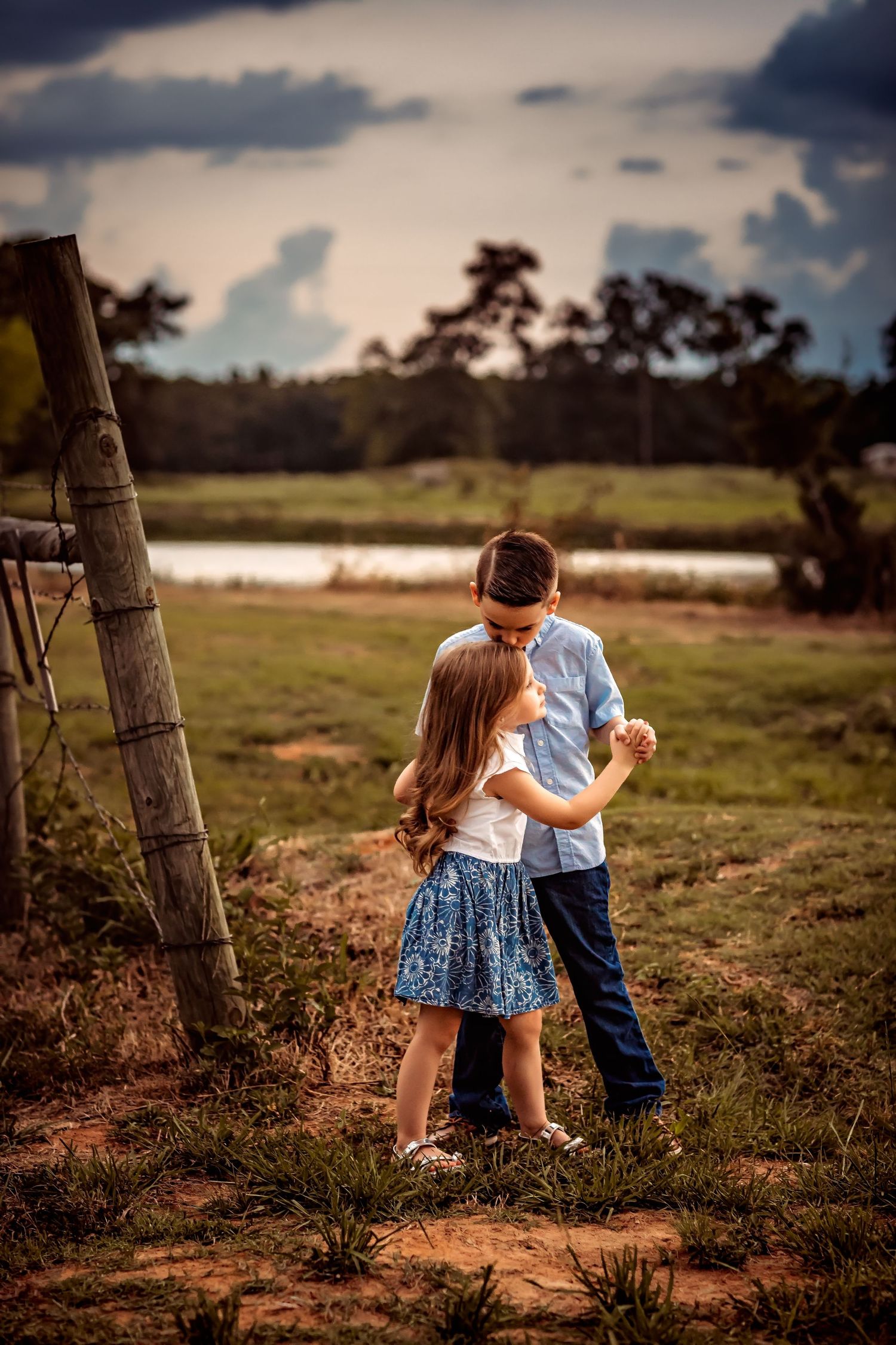 Two young children play together in a rustic countryside setting at sunset near a wooden fence post.
