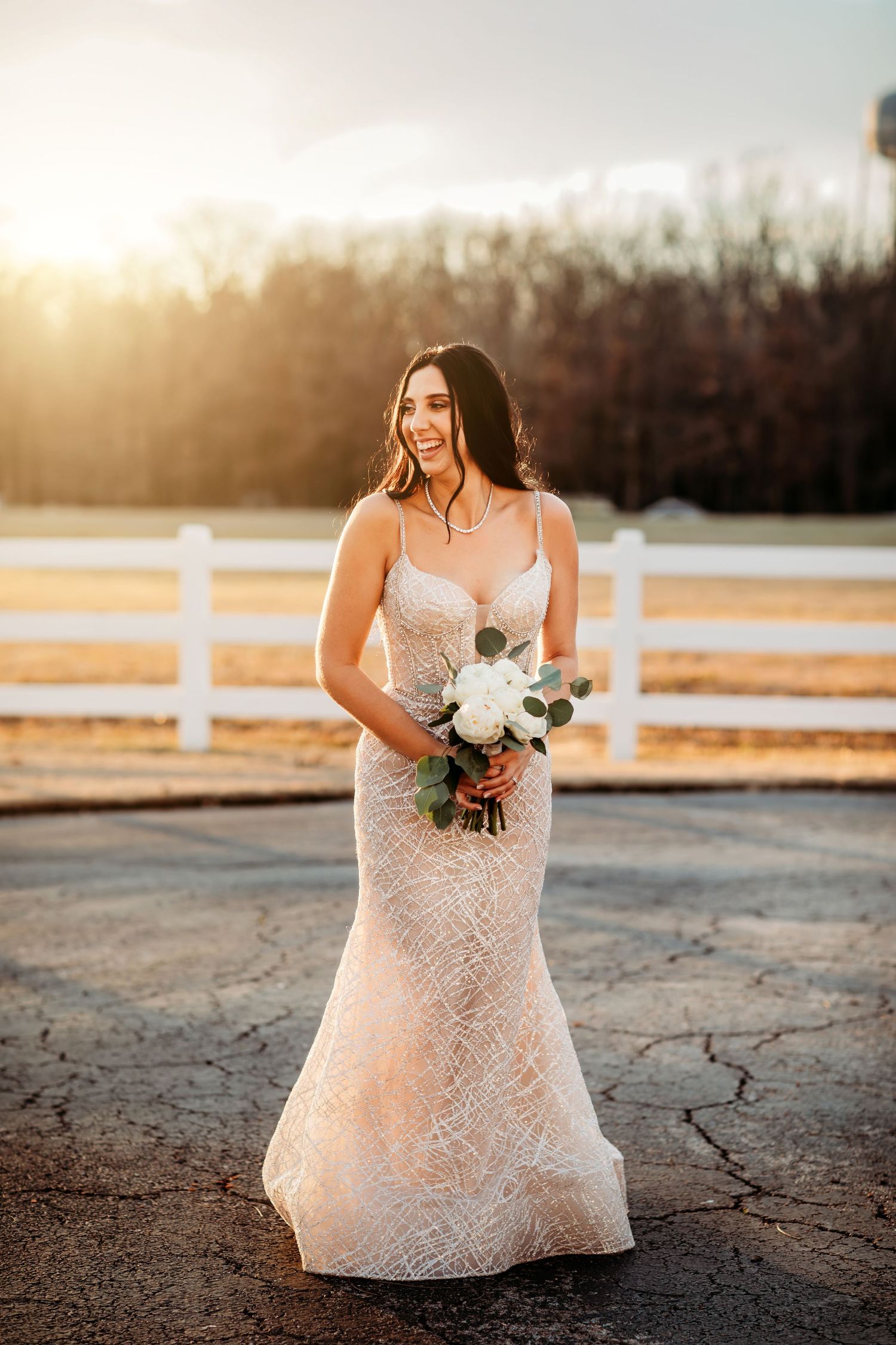 Bride in lace mermaid gown holds white bouquet at golden hour near white fence.