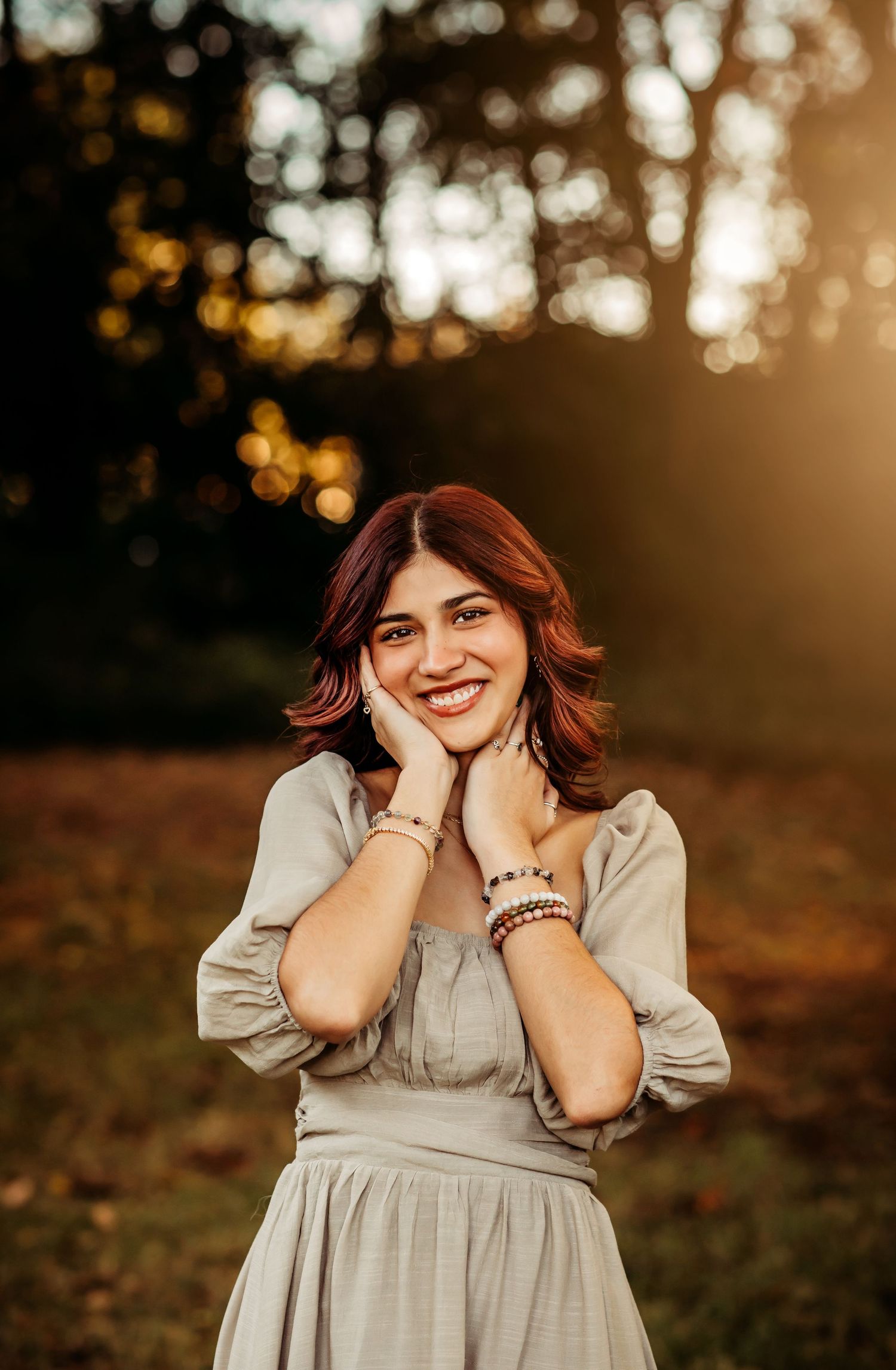Smiling young woman with red hair in a sage dress posing outdoors at golden hour with bokeh trees background.