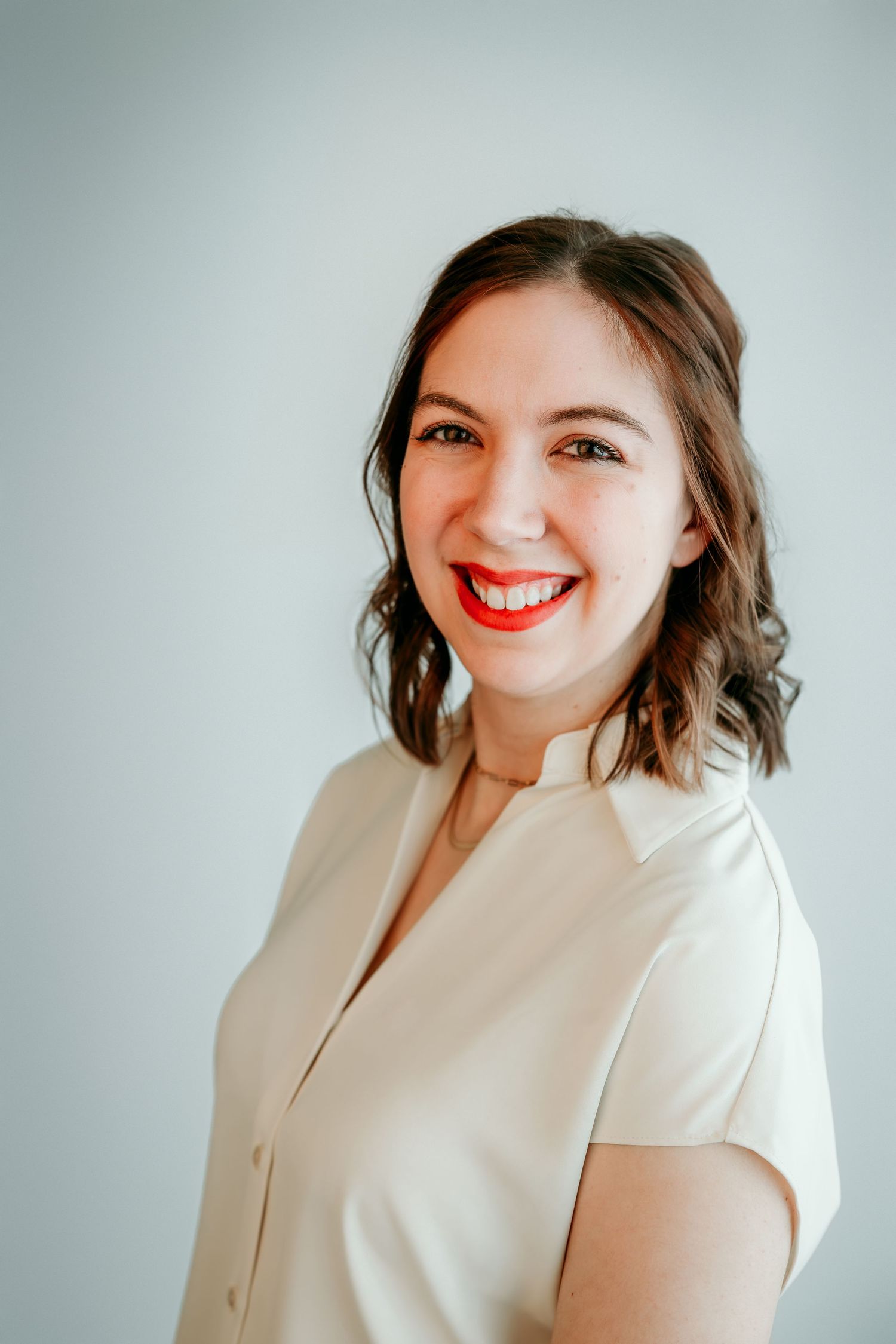 Smiling woman with red lips and brown hair wearing a cream blouse against a light gray background.