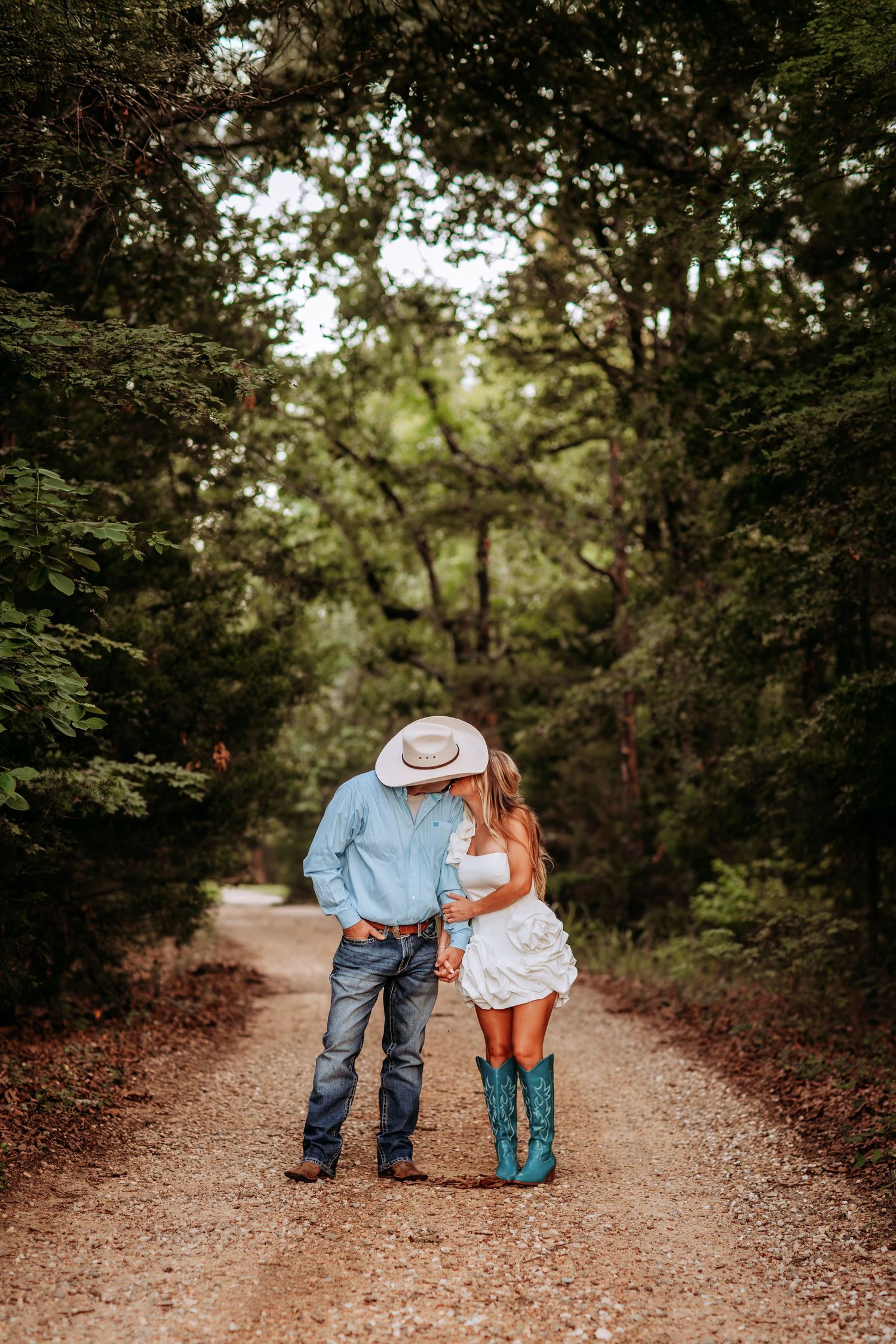 Couple in cowboy hats holding hands on a tree-lined dirt path, woman wearing white dress and turquoise boots.