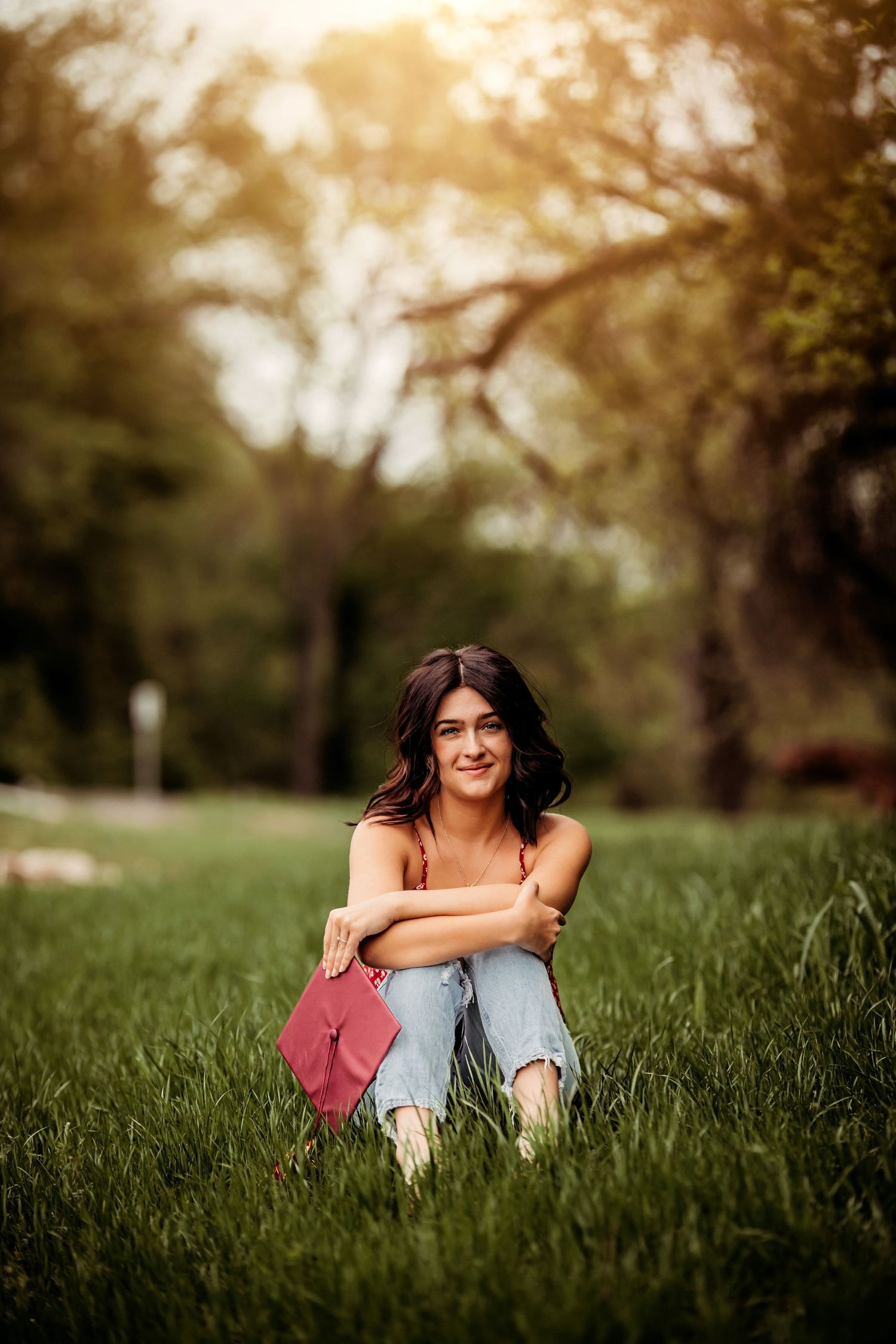 Young woman in ripped jeans sitting on green grass in a sunny park, hugging her knees and smiling.