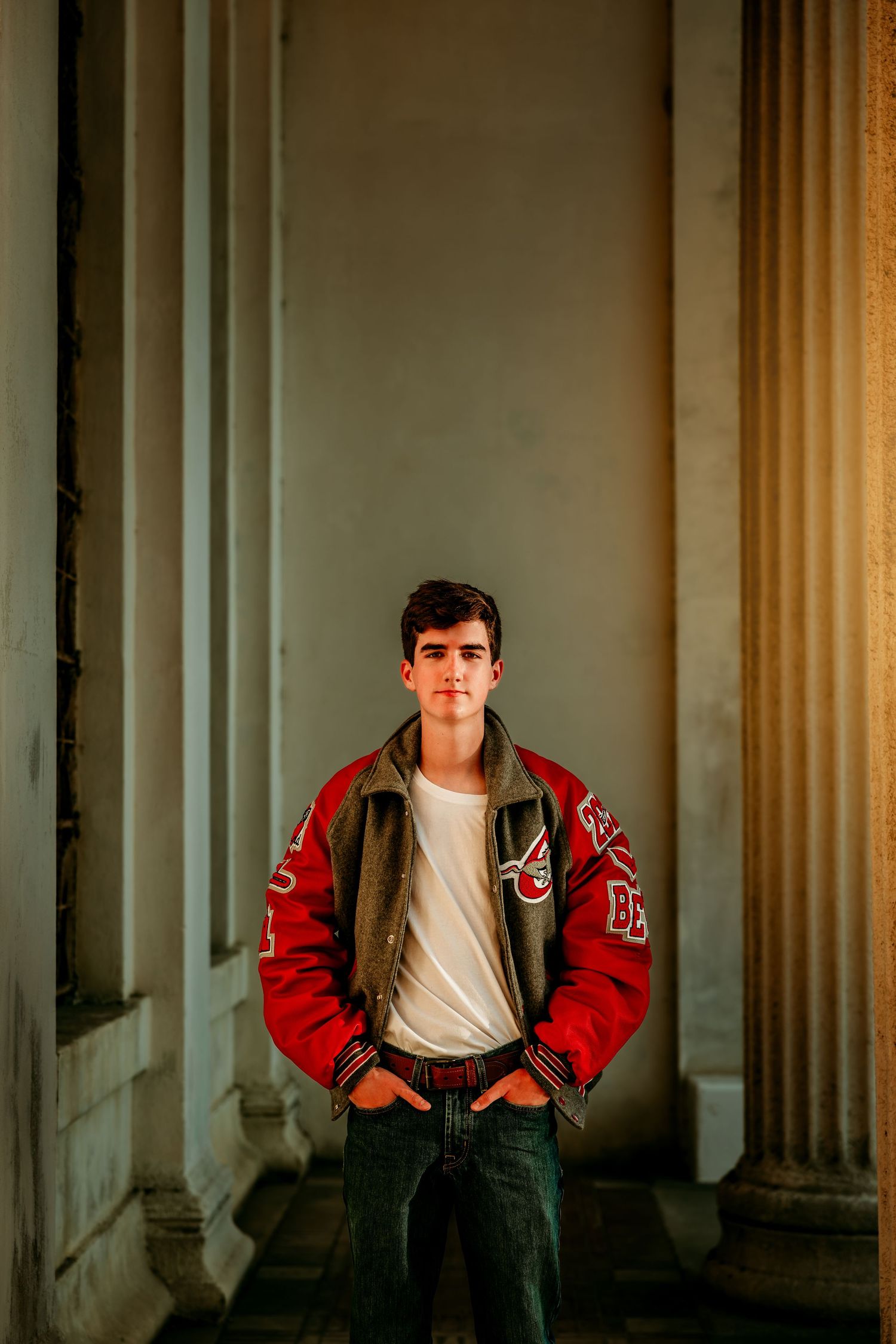 Young man in red varsity jacket stands confidently between stone columns, hands in pockets, in warm golden light.