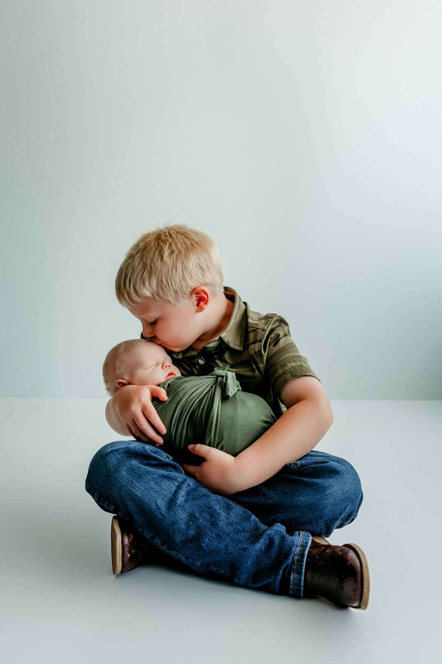 Blonde toddler boy sitting cross-legged, gently kissing a swaddled newborn baby wrapped in green cloth.