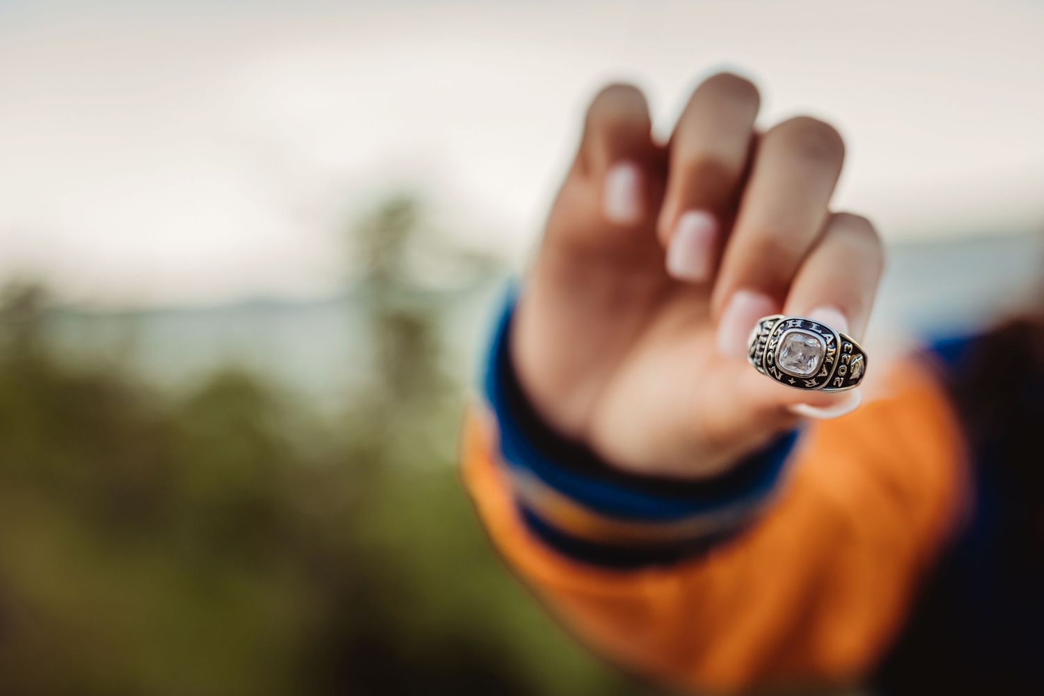 Person wearing an orange jacket holds up a championship ring toward the camera with a blurred green landscape background.