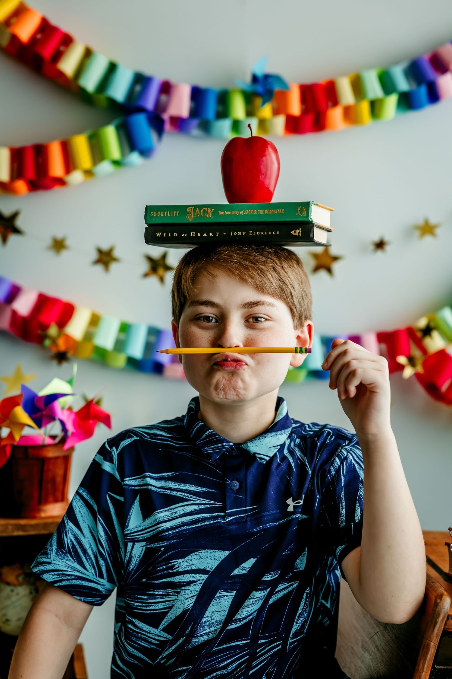 Boy balancing books and apple on head while holding pencil as mustache, with colorful rainbow decorations behind him.