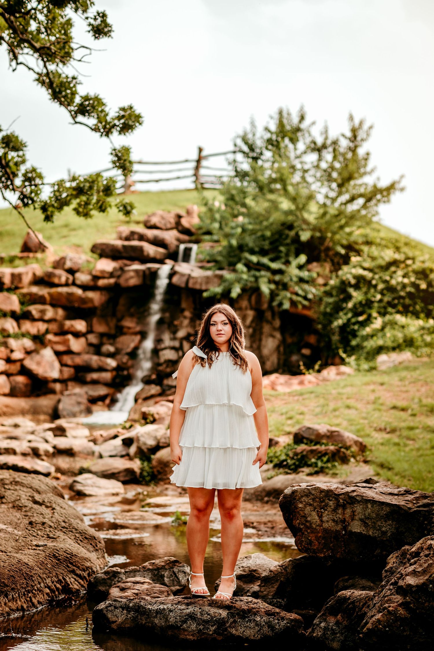 Young woman in white tiered dress standing on rocks near a small waterfall in a lush green outdoor setting.