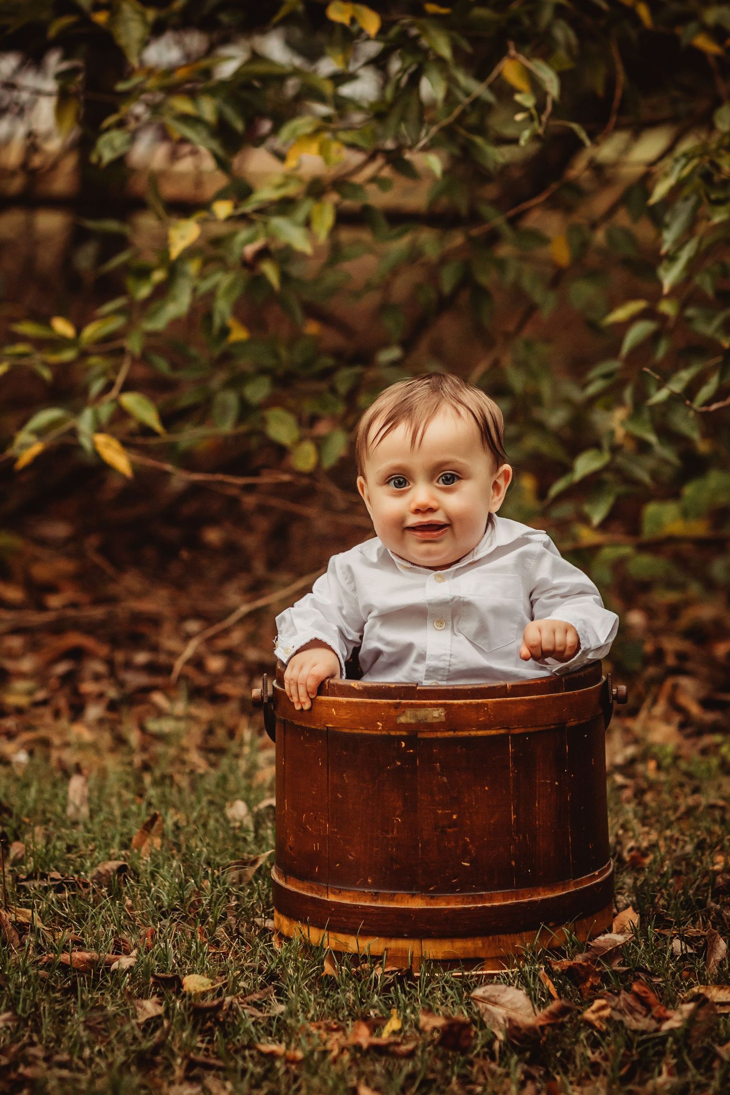 A small child sits in a vintage wooden barrel outdoors among fallen autumn leaves.