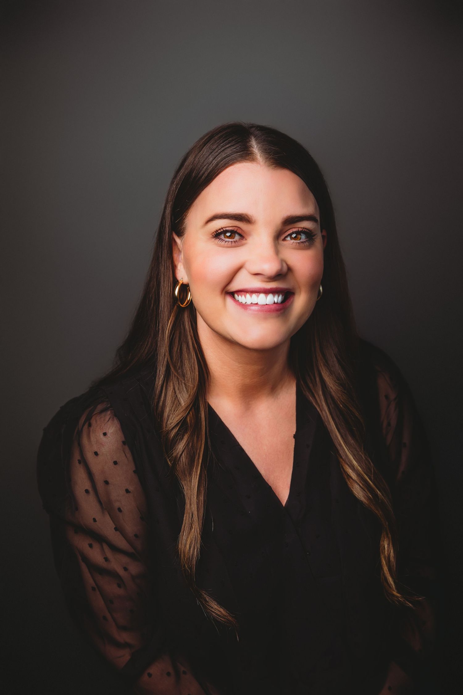Smiling young woman with long brown hair wearing a black blouse and gold hoop earrings against a dark background.