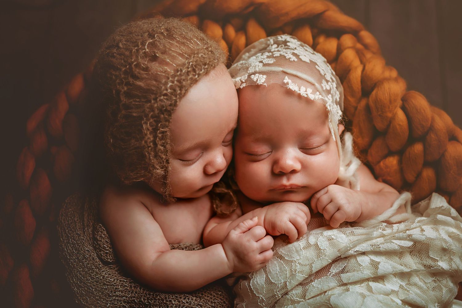 Two sleeping newborn twins in a cozy basket, one wearing a knit bonnet and the other a lace headband.
