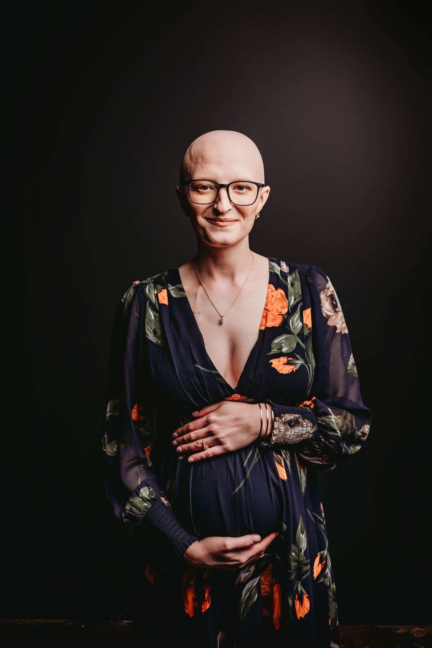 Smiling pregnant woman with shaved head wearing floral dress, posing against dark background.