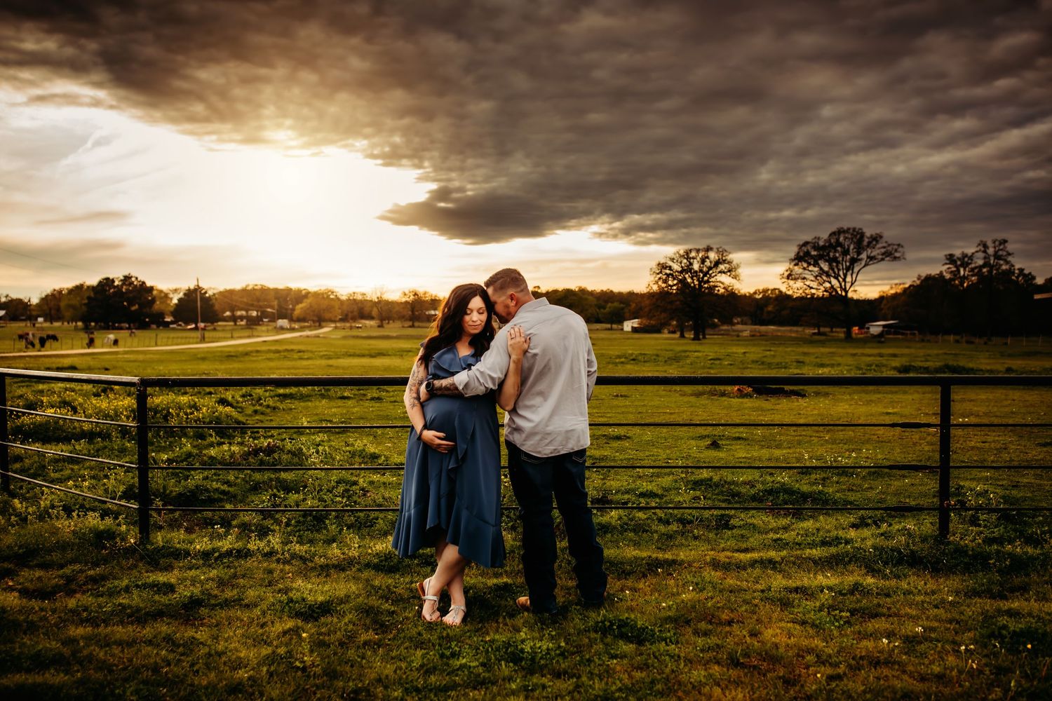 Couple embracing at golden hour in a green field with dramatic storm clouds and wooden fence backdrop.