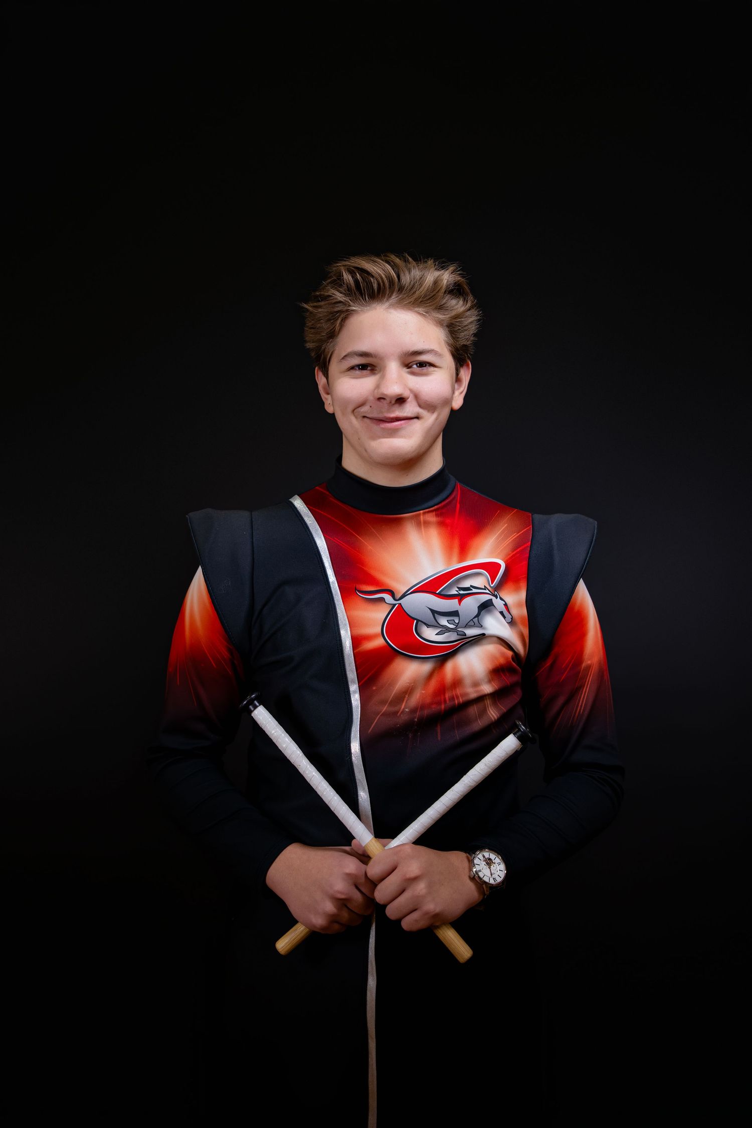Young drummer in red and black marching band uniform holds drumsticks, smiling against dark background.