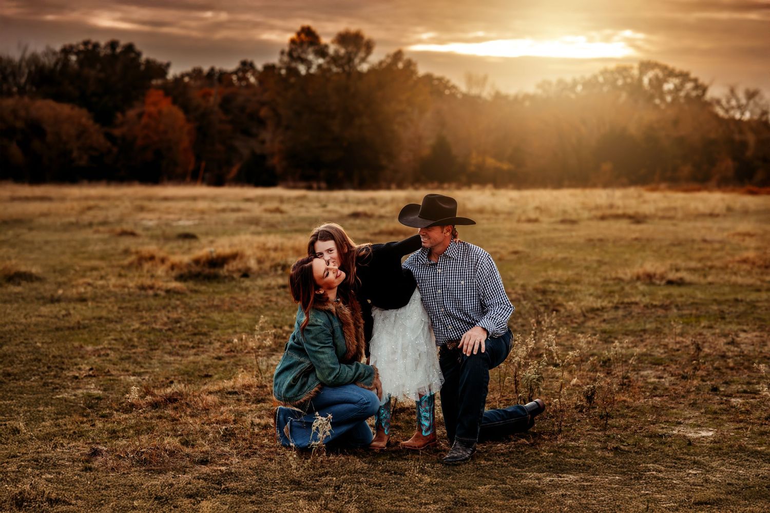 Family of three poses in golden sunset light in an autumn field, parents kneeling beside child in white dress.