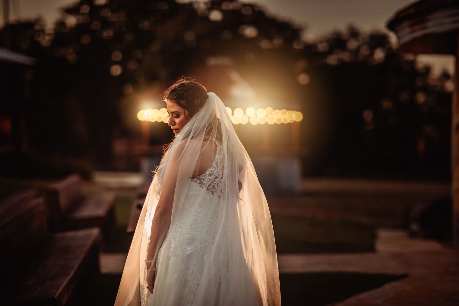 Bride in lace wedding gown and long veil poses outdoors at dusk with warm glowing lights in background.