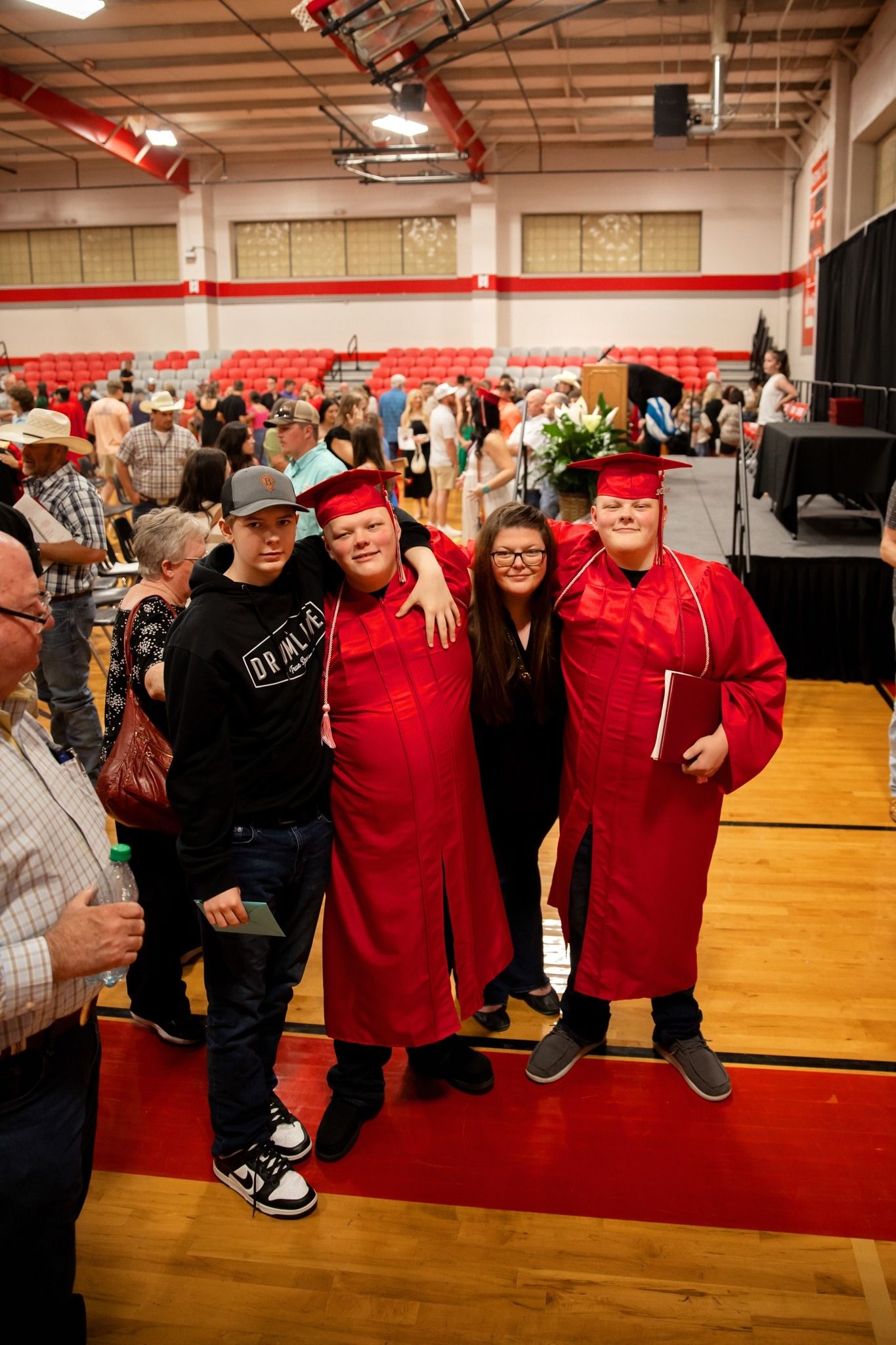 A group of students and friends pose together in red graduation gowns during a graduation ceremony in a school gymnasium.