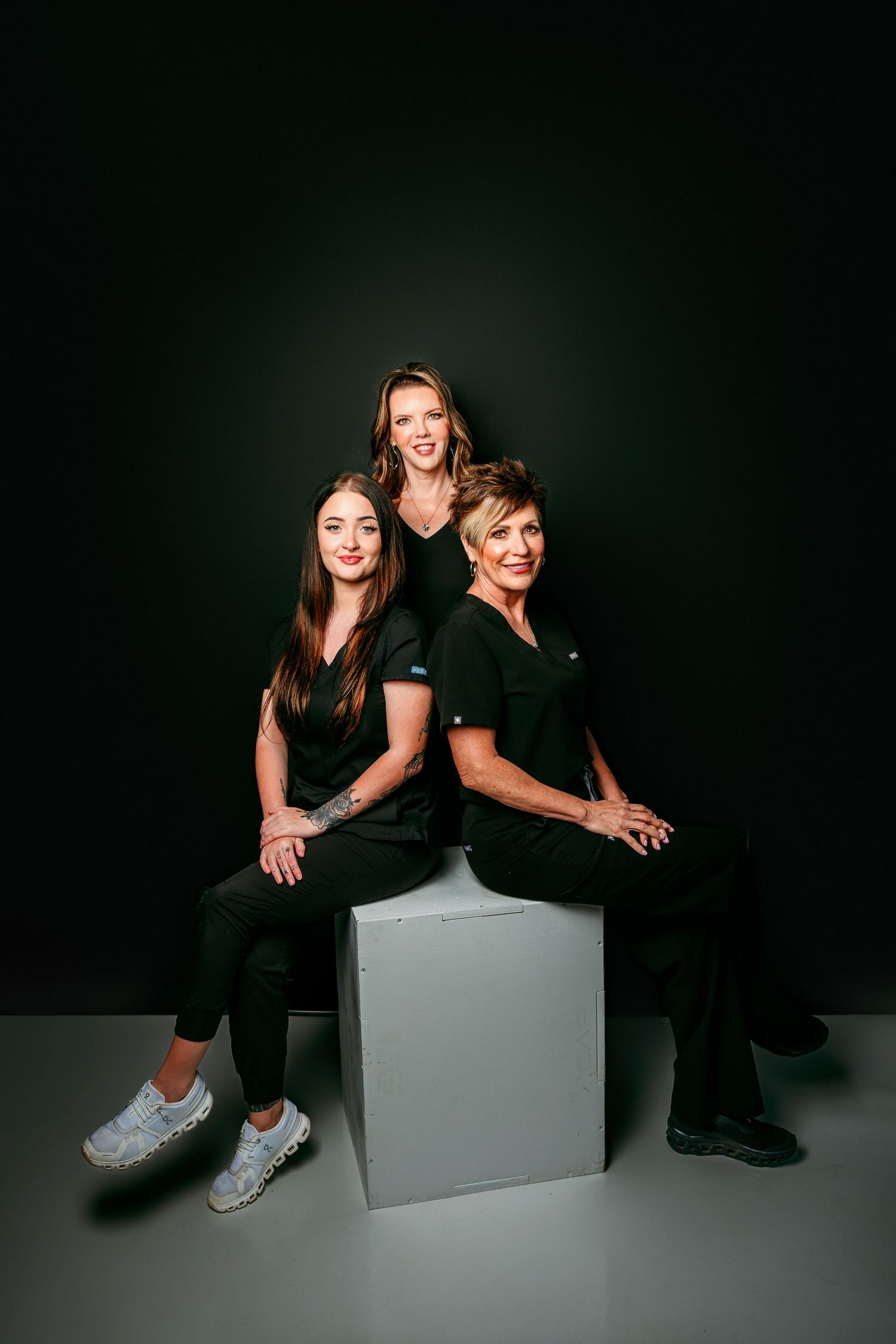 Three women in black scrubs pose together in a professional studio portrait against a dark background.