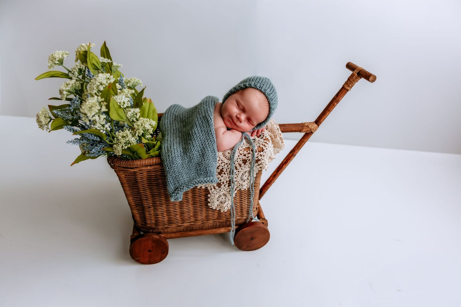Sleeping newborn baby in blue knit bonnet nestled in rustic wicker wagon with white flowers.