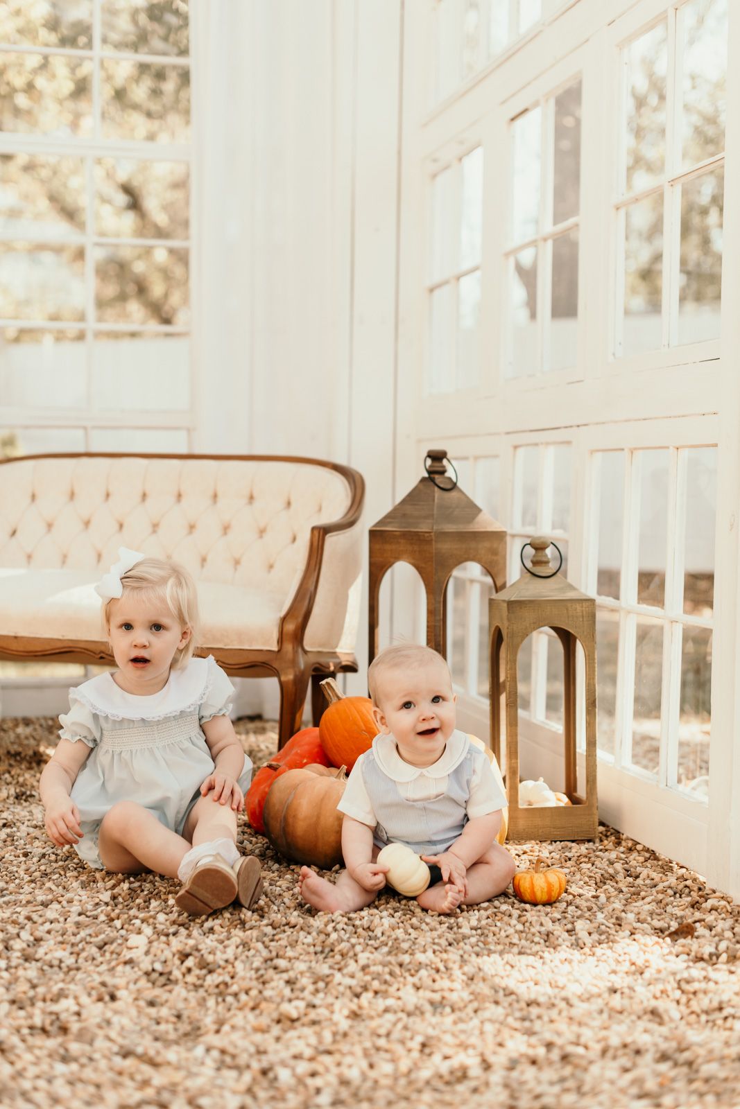 Two babies sitting on carpet with autumn pumpkins and decorative lanterns in a bright sunlit room.