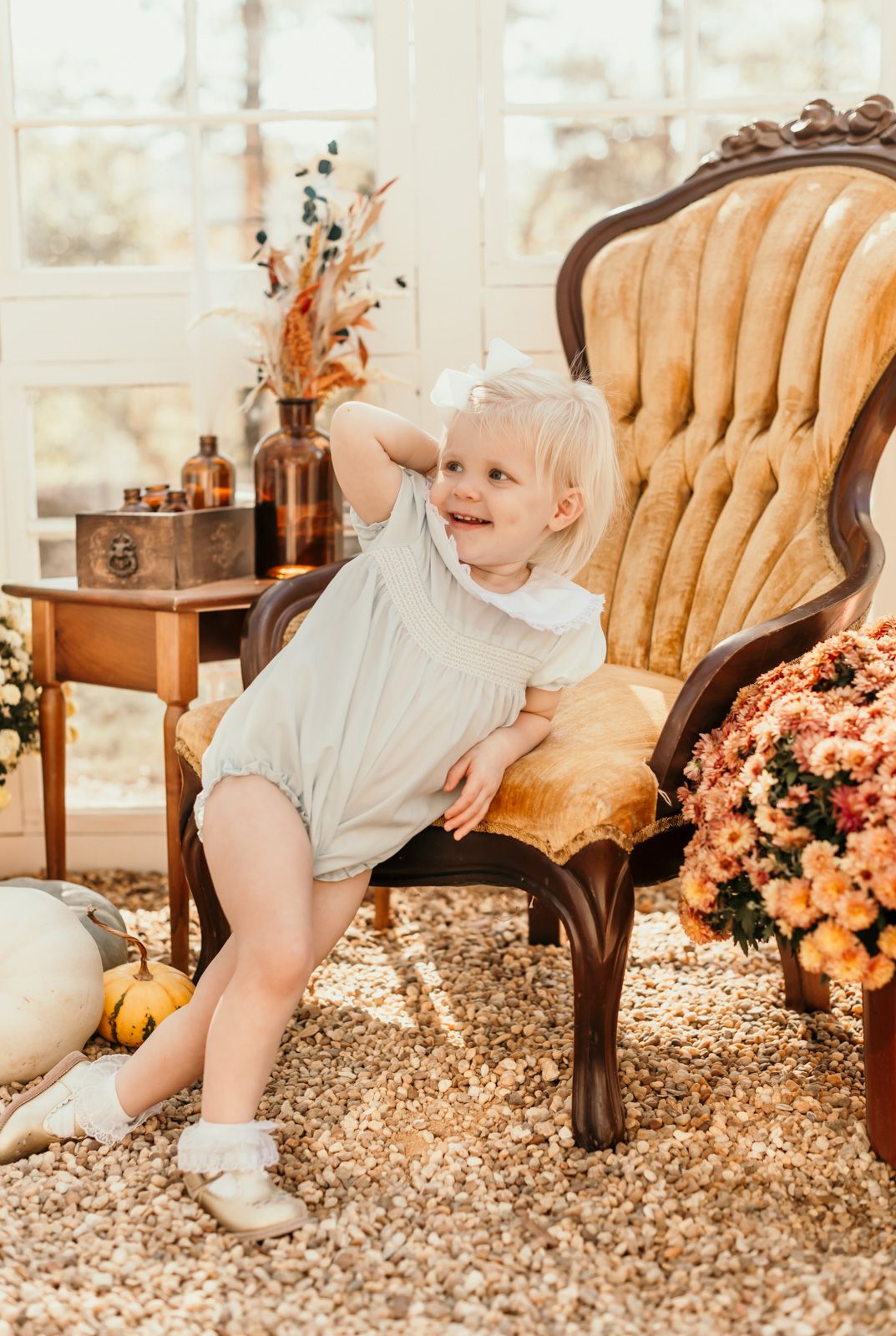 A vintage tufted beige chair sits beside autumn decor and flowers in a bright sunlit room.