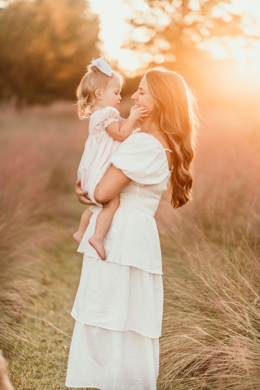 Mother and daughter in white dresses embrace in a sunlit field of tall grass during golden hour.
