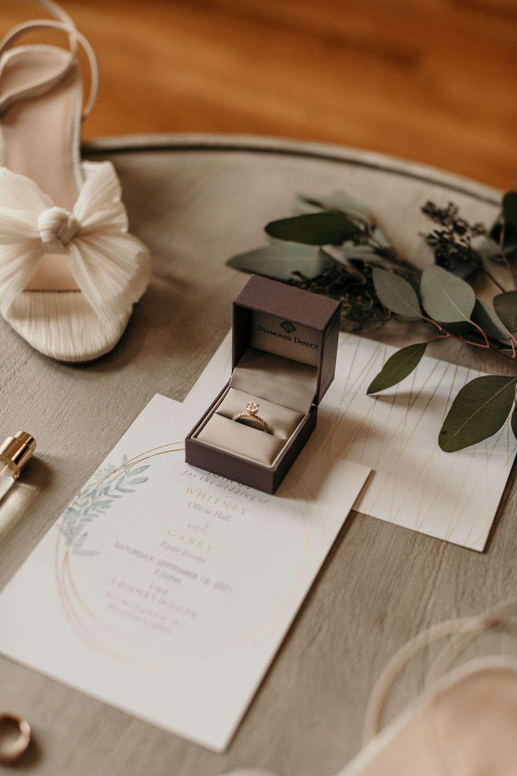Wedding details flatlay with white stationery, floral elements, and bridal accessories on a light wooden surface.