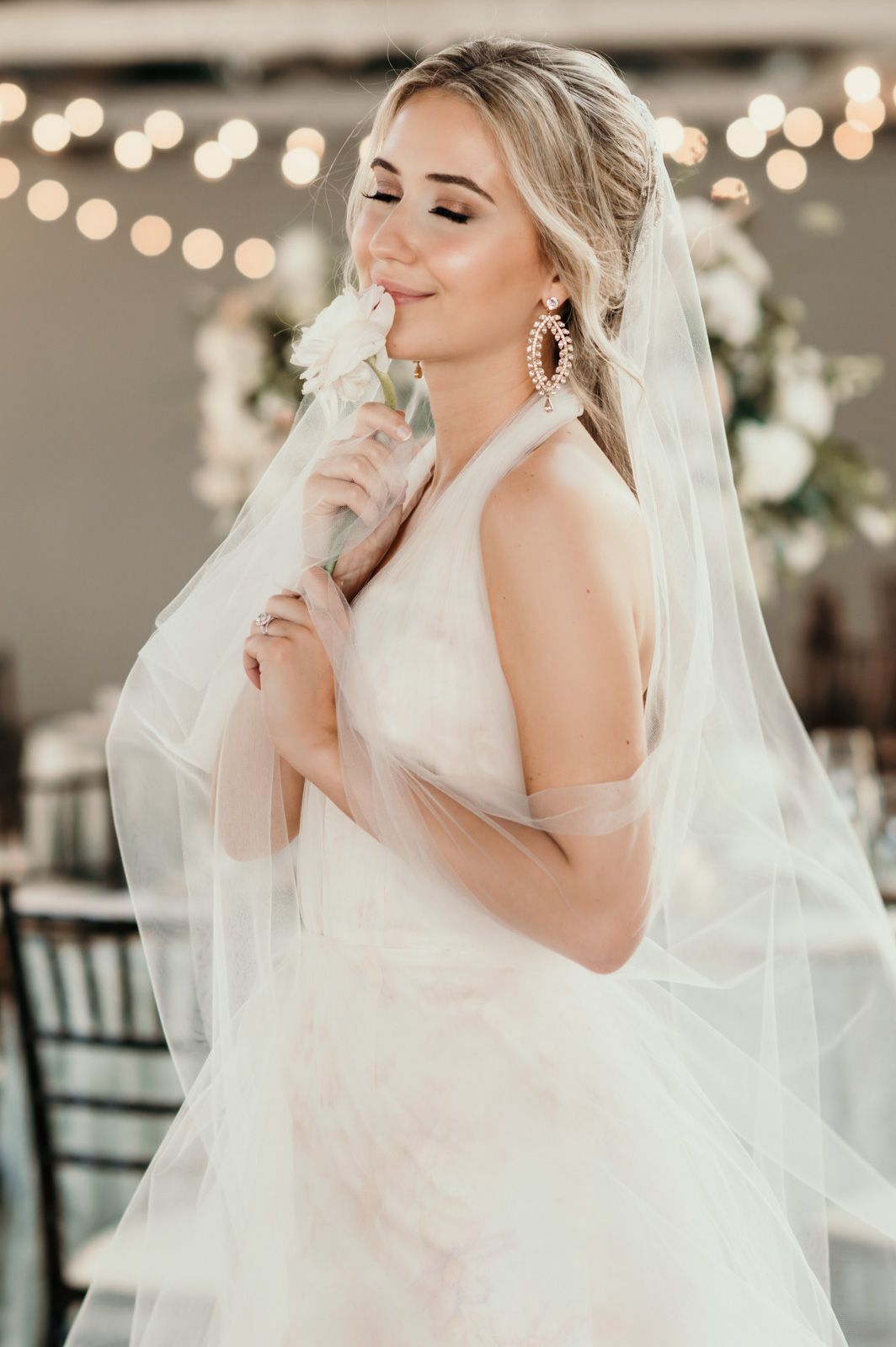 A bride in an elegant white wedding gown poses with flowing veil and sparkly earrings against a bokeh light background.