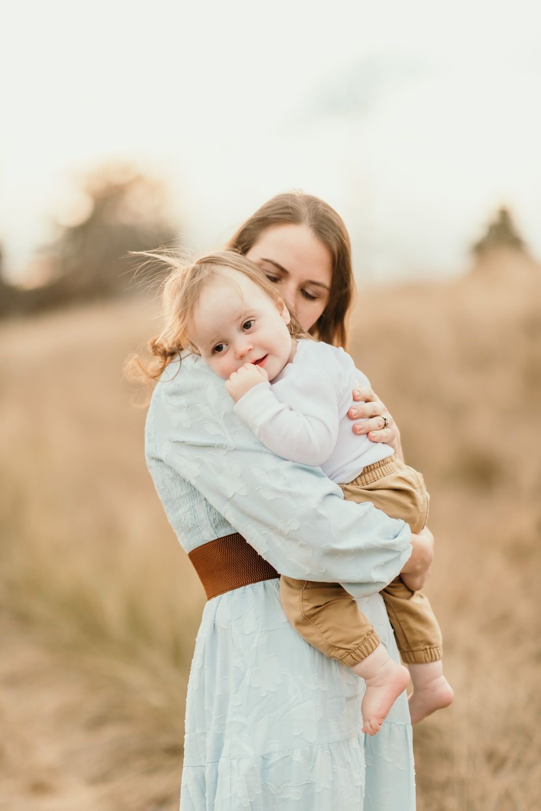 A tender moment captured in a field as a parent embraces a small child wearing a light blue dress at sunset.