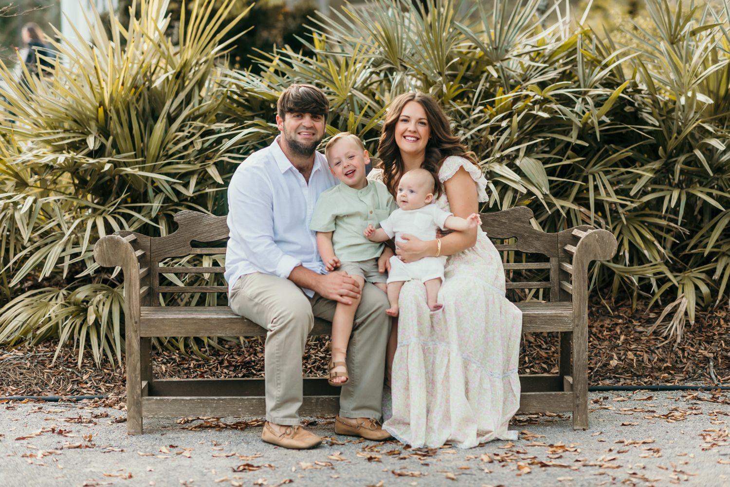 Family in white outfits sits together on stone steps surrounded by tropical plants for outdoor photo session.