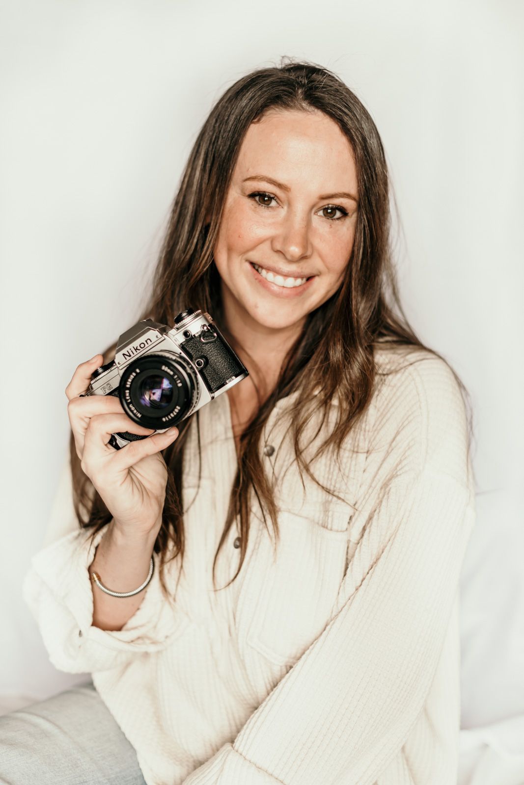 A photographer in a cream-colored sweater holds a vintage film camera while smiling against a light background.