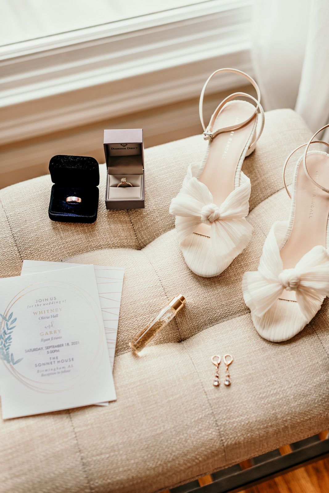 Wedding accessories including rings, invitation, shoes, and jewelry arranged on a beige textured surface in natural light.