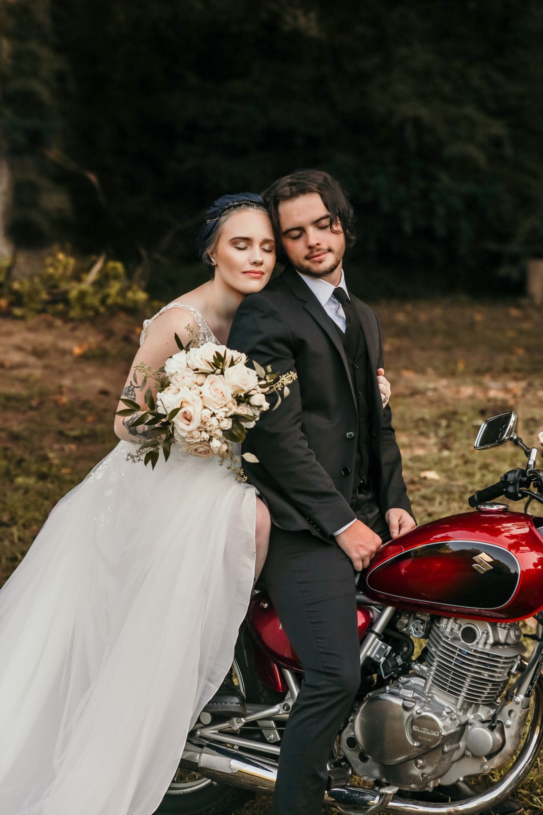 A romantic wedding photo featuring a couple posing with a vintage red motorcycle in an outdoor setting.